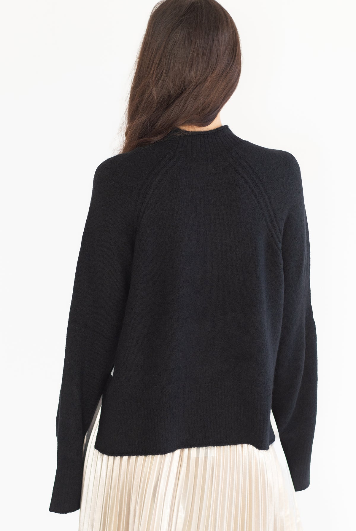 A woman shown from the back wearing a black knit sweater and a cream pleated skirt against a plain white background.