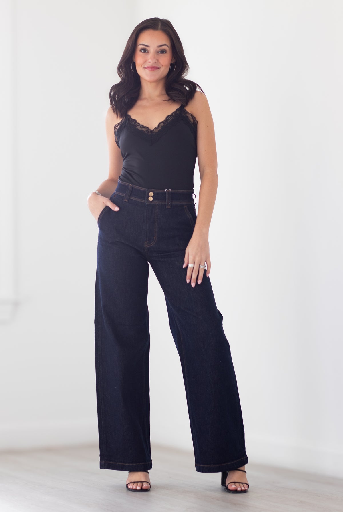 A woman modeling a black lace-trimmed camisole top paired with dark wide-leg denim jeans and black sandals, standing against a bright white wall.