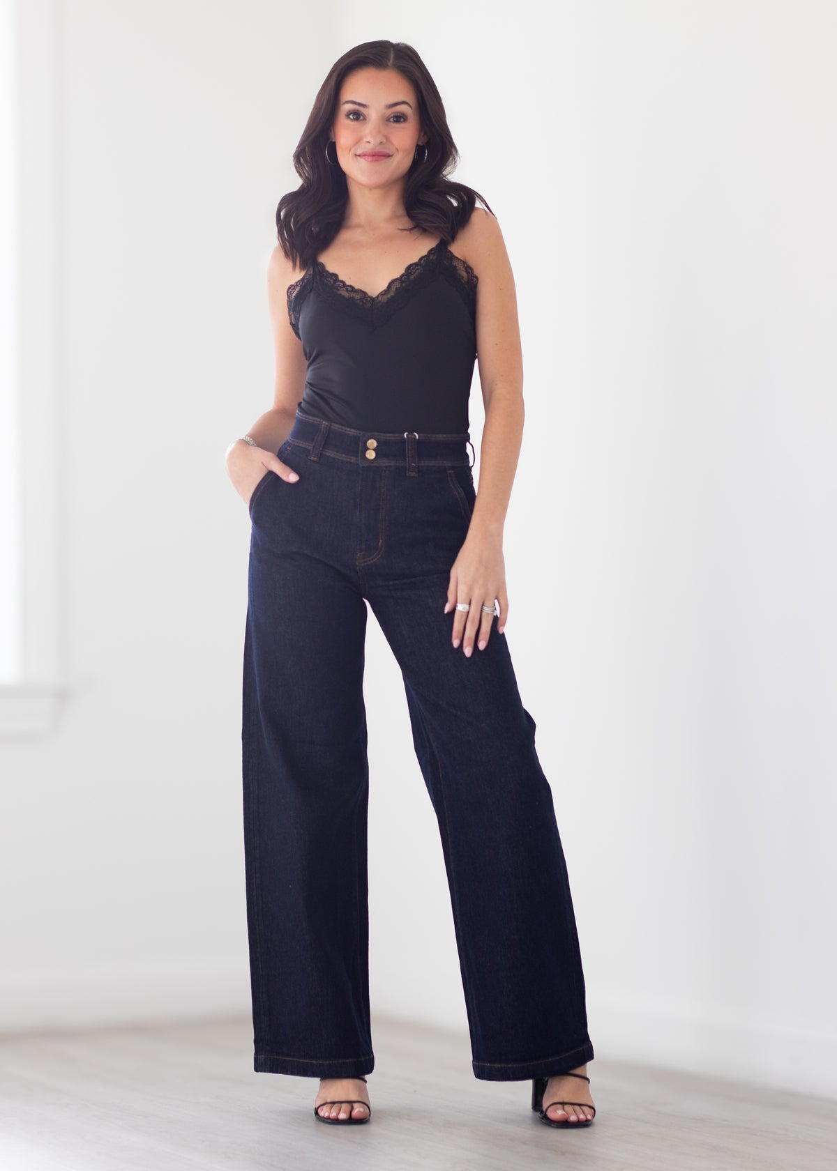 A woman modeling a black lace-trimmed camisole top paired with dark wide-leg denim jeans and black sandals, standing against a bright white wall.