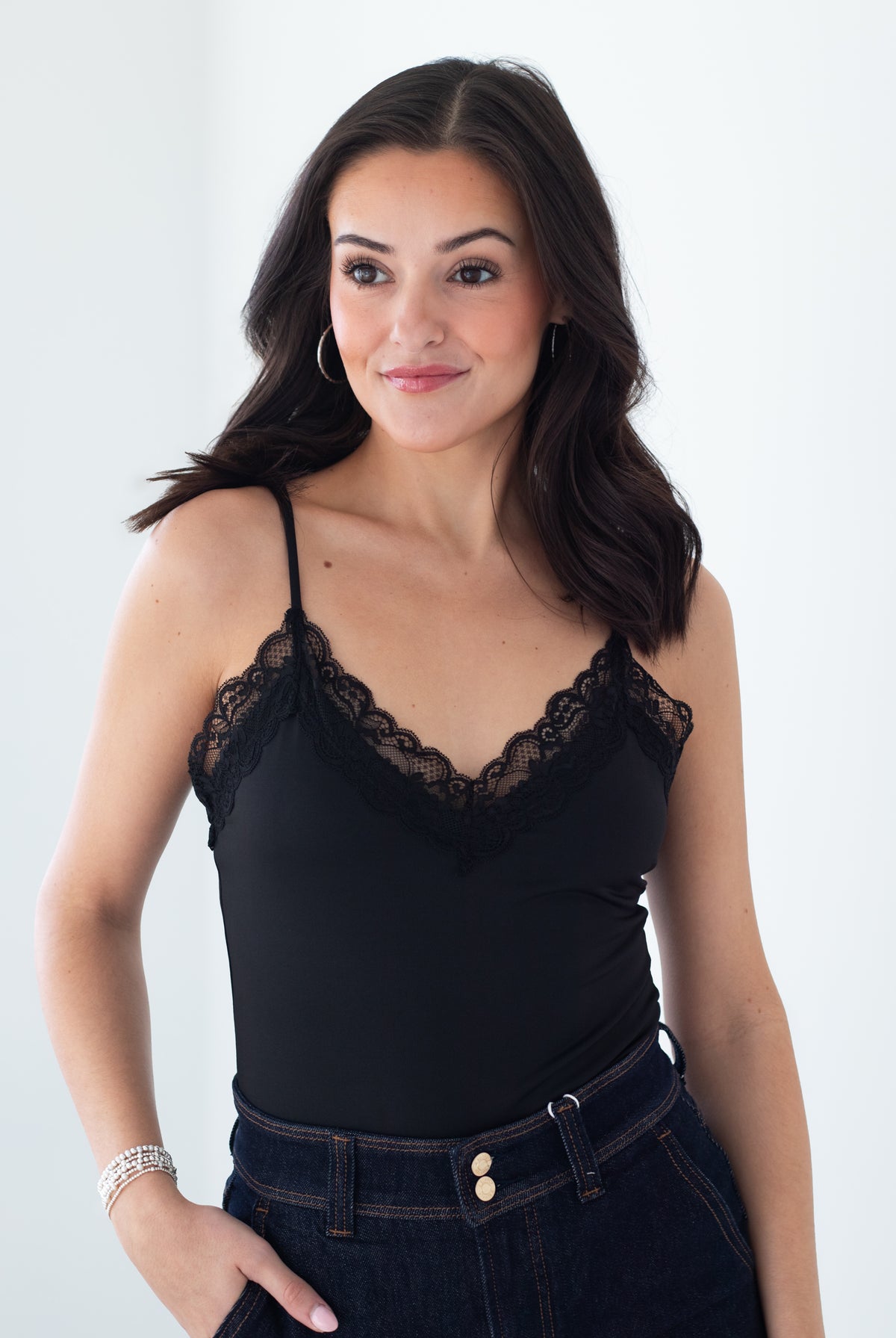 A woman wearing a black camisole top with delicate lace trim along the straps and neckline, paired with dark blue jeans, set against a plain white studio background.