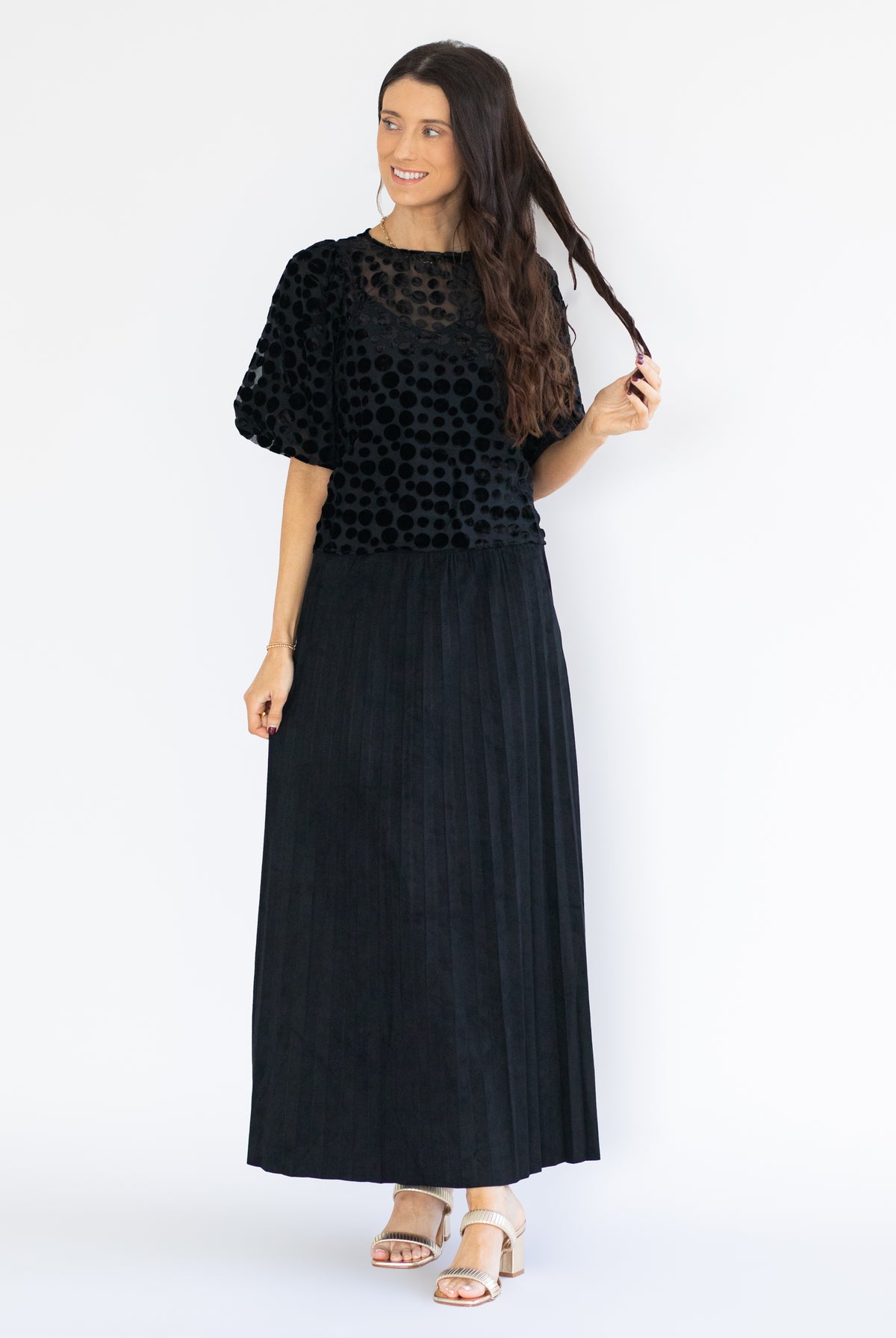 A woman modeling a black two-piece outfit consisting of a short-sleeve top with a textured, dotted pattern and a matching long pleated skirt, paired with beige sandals against a white studio background.