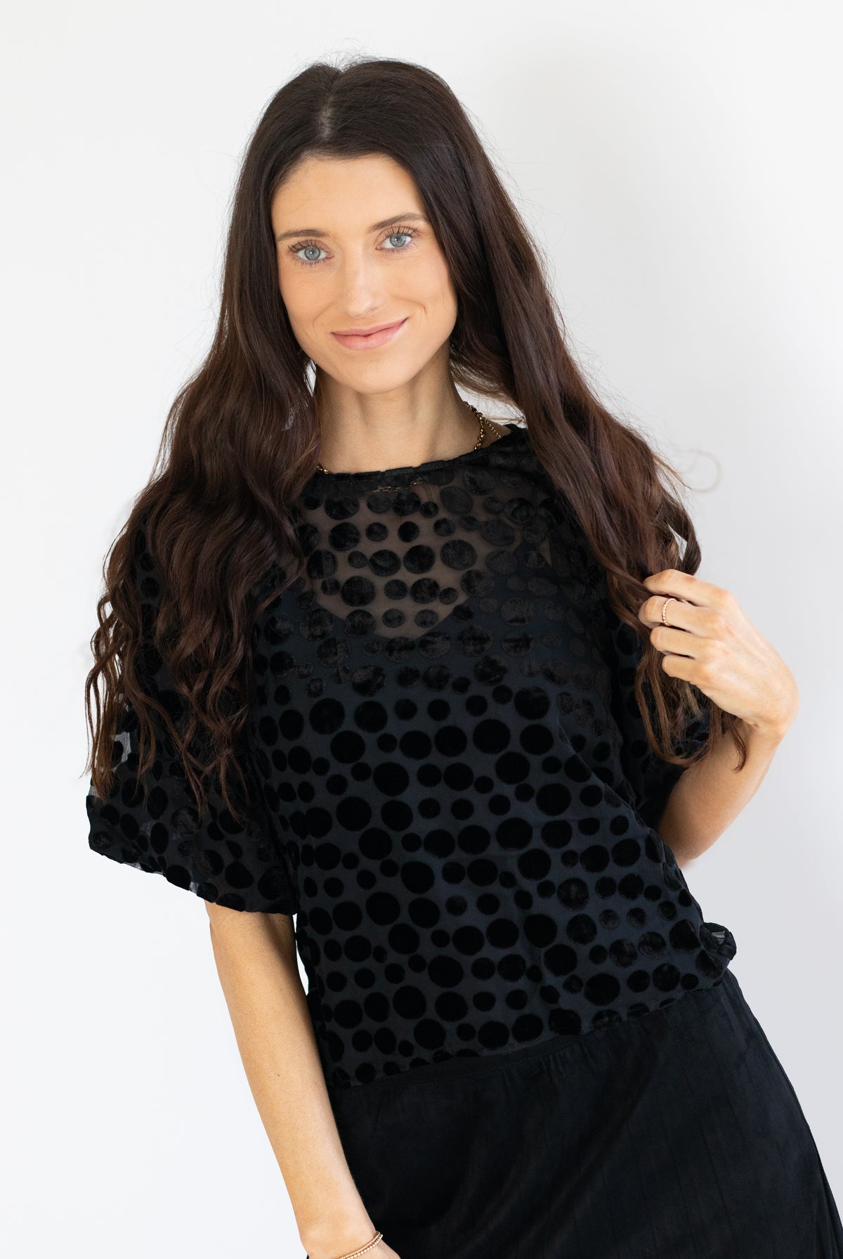 A woman with long, wavy brown hair wearing a black short-sleeve top with a textured polka dot pattern and a sheer mesh neckline, paired with a black skirt, set against a plain white background.