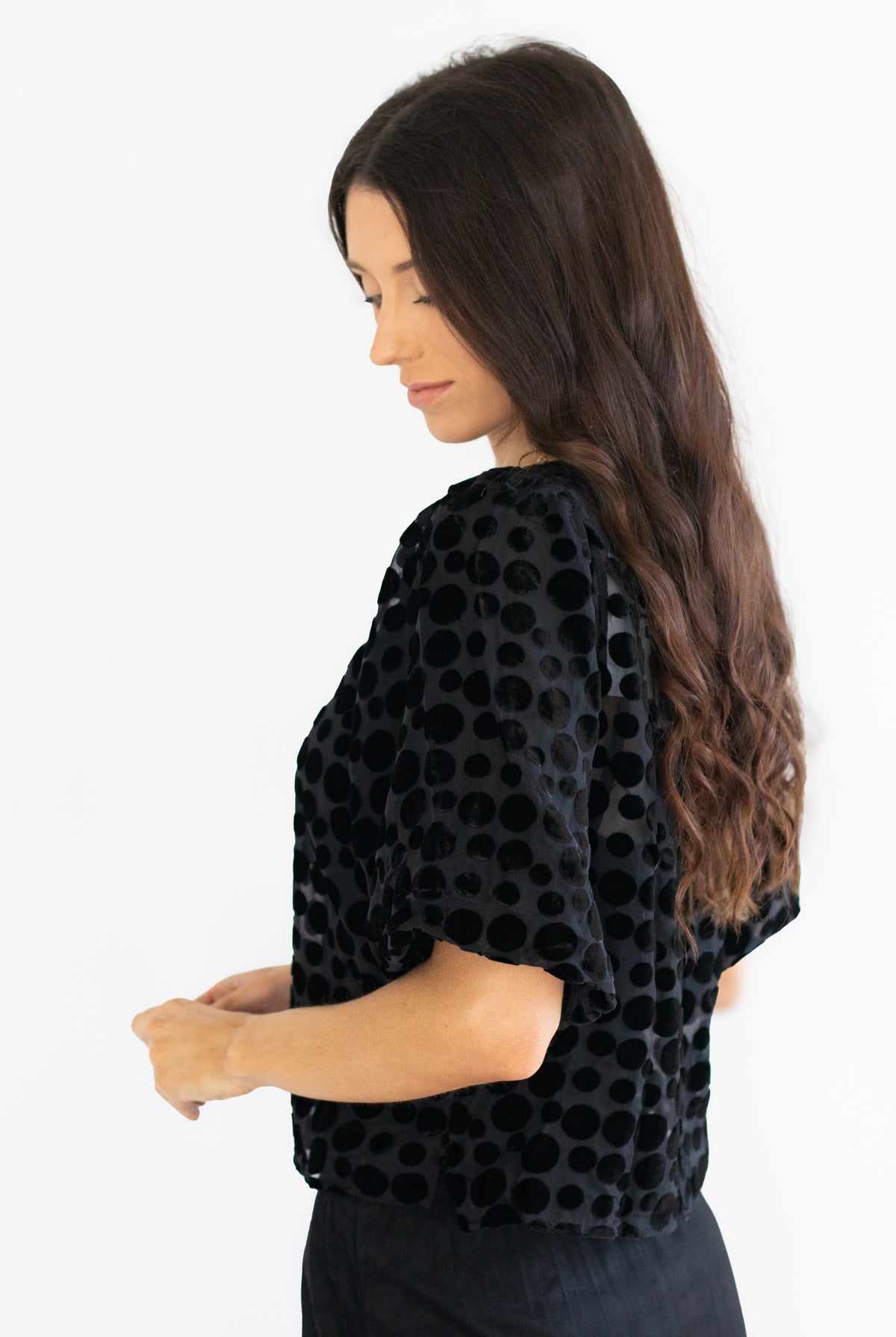 A woman with long dark brown hair is shown from a side-back angle wearing a black velvet top with a circular polka dot pattern and short sleeves, paired with black pants, against a plain white background.
