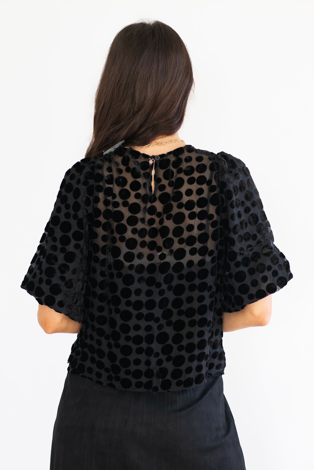 A woman viewed from the back wearing a black sheer mesh top with a black polka dot pattern and puff sleeves, paired with a black skirt, against a plain white background.