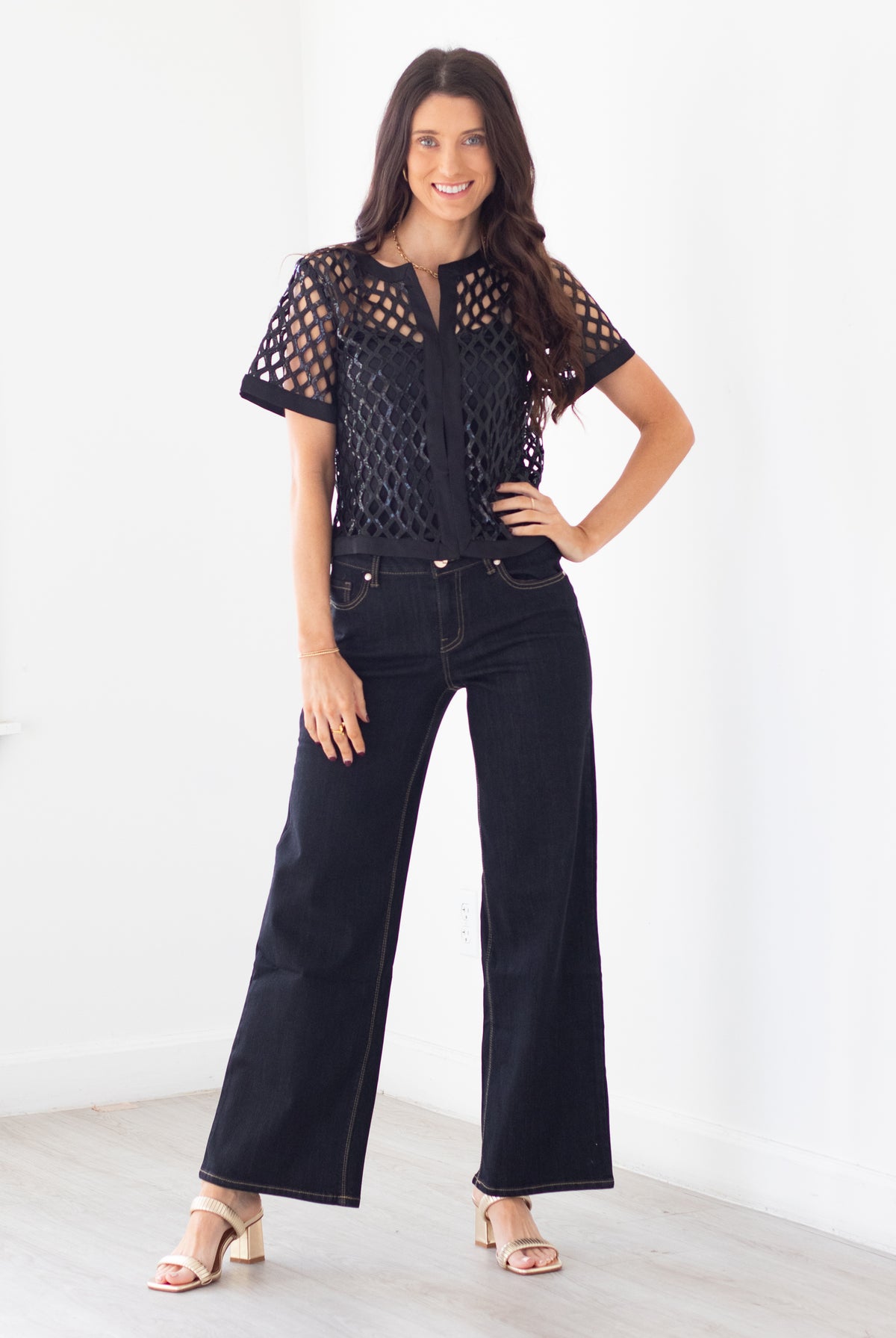 A woman modeling a black short-sleeve crochet top with sheer eyelet sleeves and dark wide-leg jeans, standing in a bright white room.