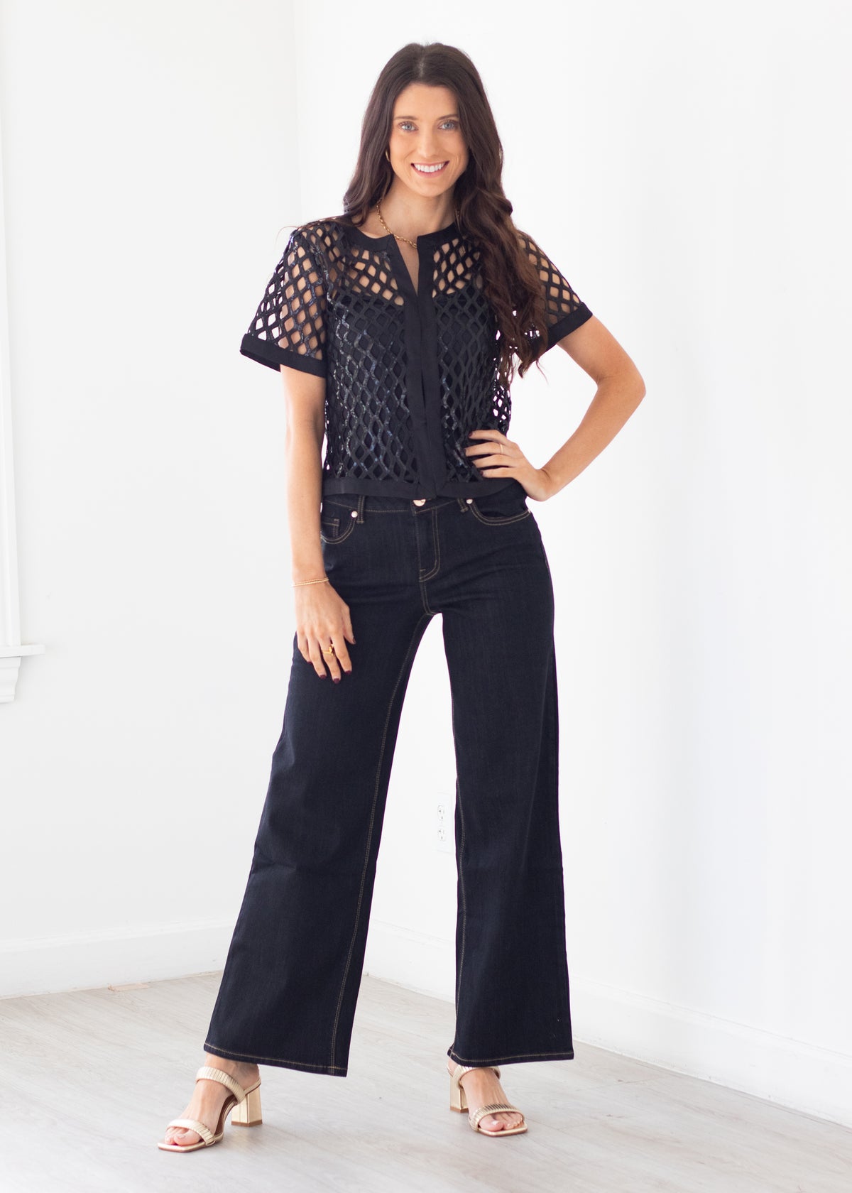 A woman modeling a black short-sleeve crochet top with sheer eyelet sleeves and dark wide-leg jeans, standing in a bright white room.