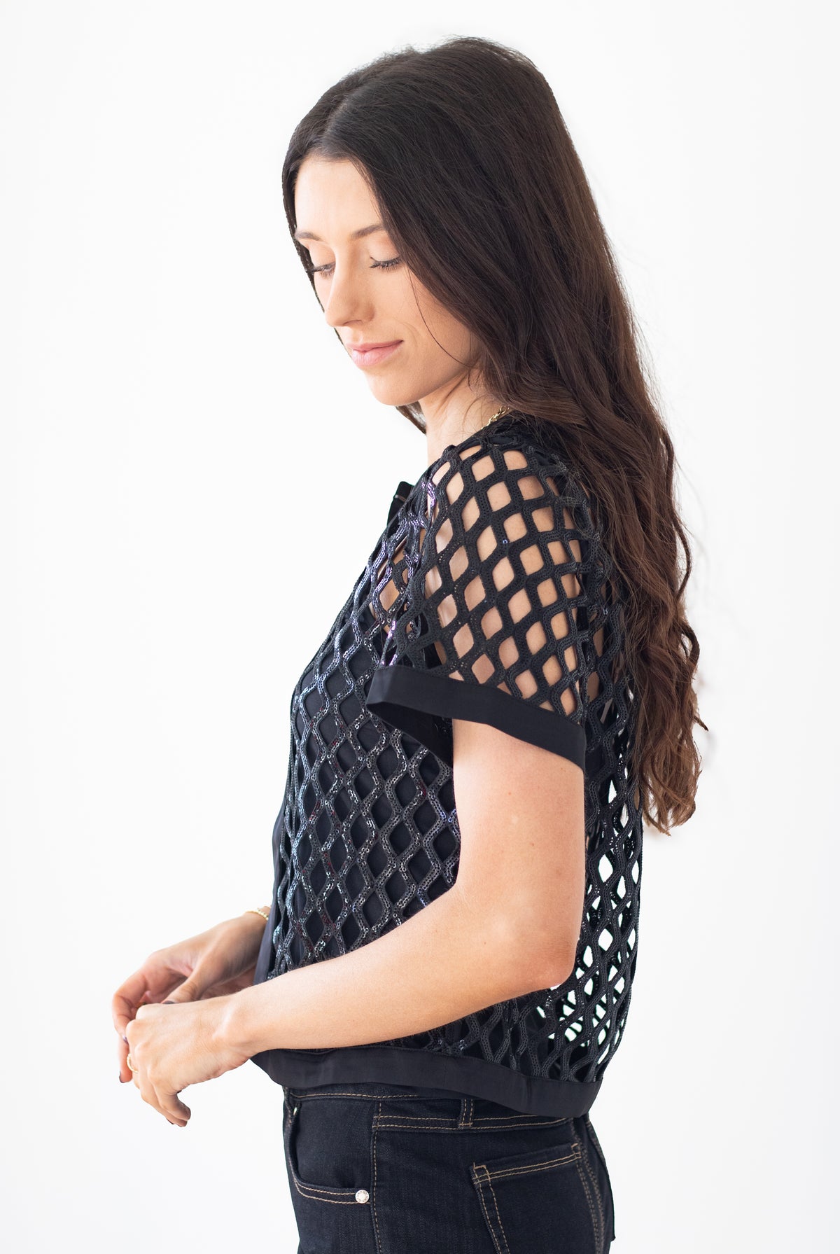 A woman with long dark hair is shown in profile wearing a black short-sleeve top with a geometric openwork lattice pattern, paired with dark jeans, against a plain white studio background.