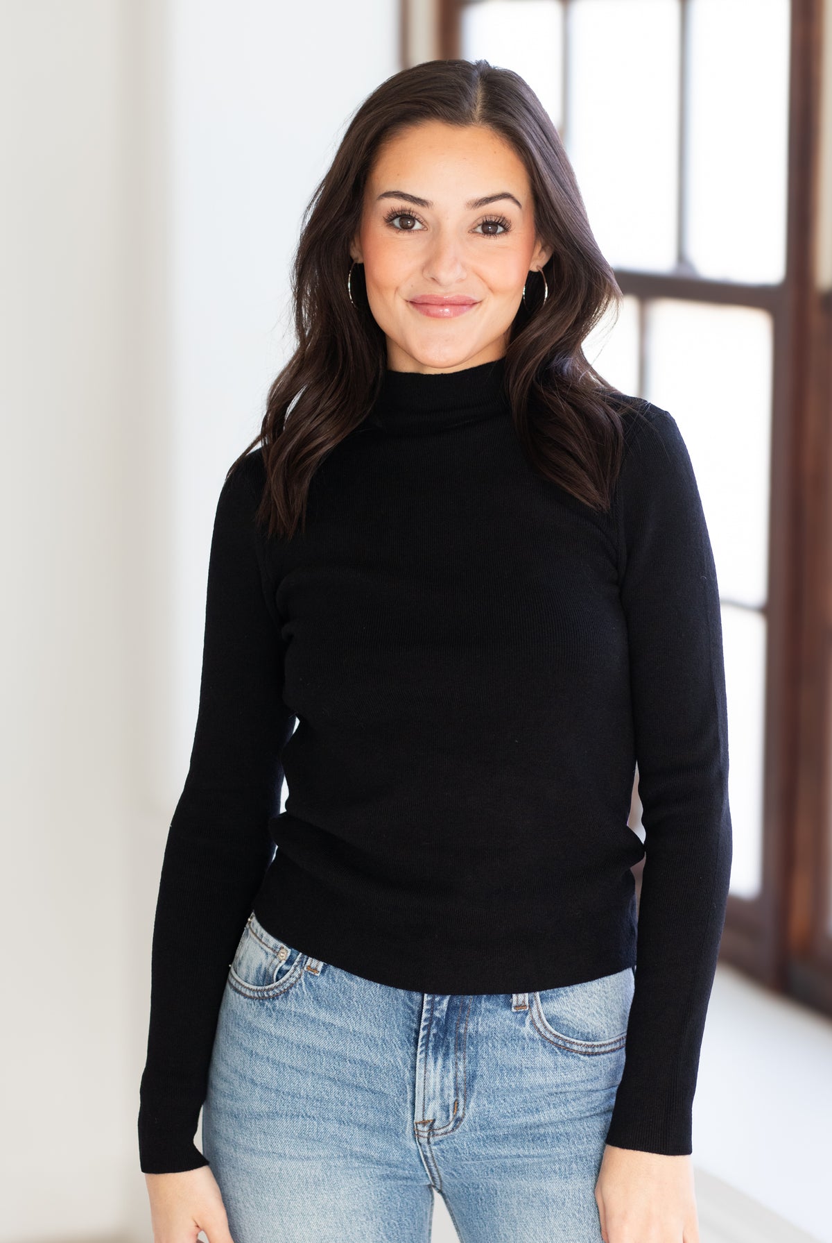 A woman with long dark hair wearing a black long-sleeve mock neck sweater and light blue jeans, standing in a brightly lit room with a window in the background.