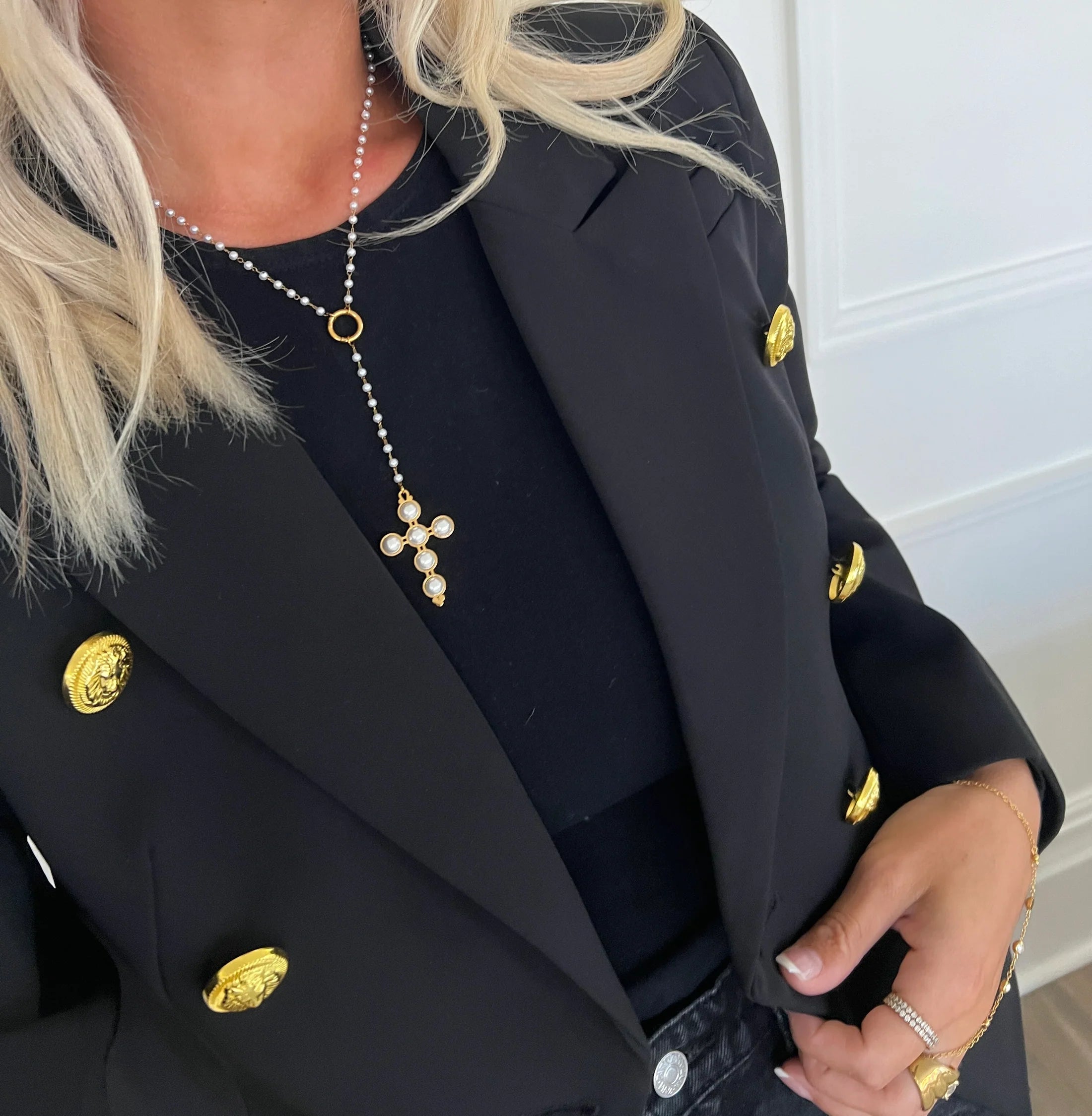 A person wearing a black jacket and a lariat pearl cross necklace with gold plating.