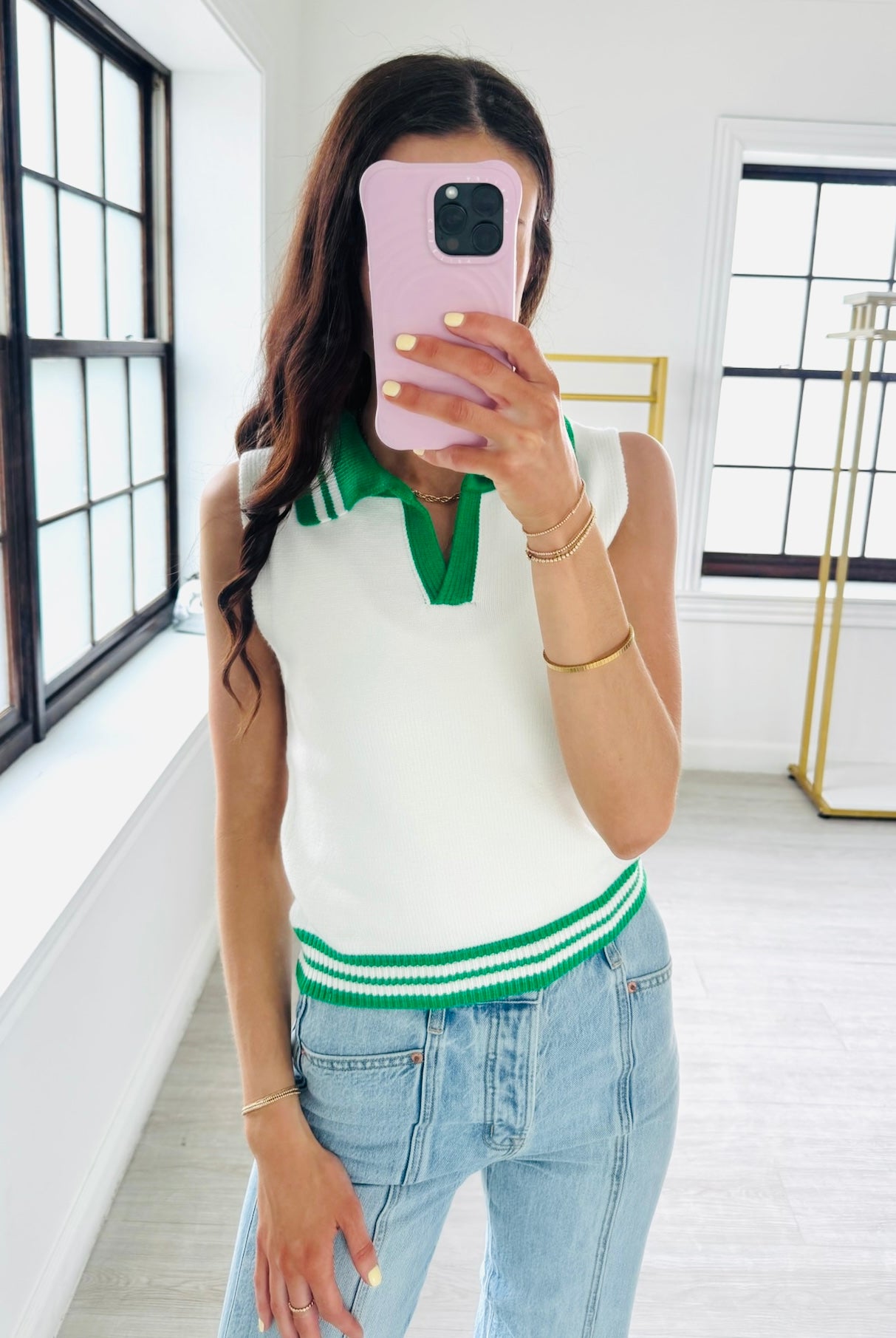 Woman taking a mirror selfie wearing a white sleeveless sweater with green trim and blue jeans.