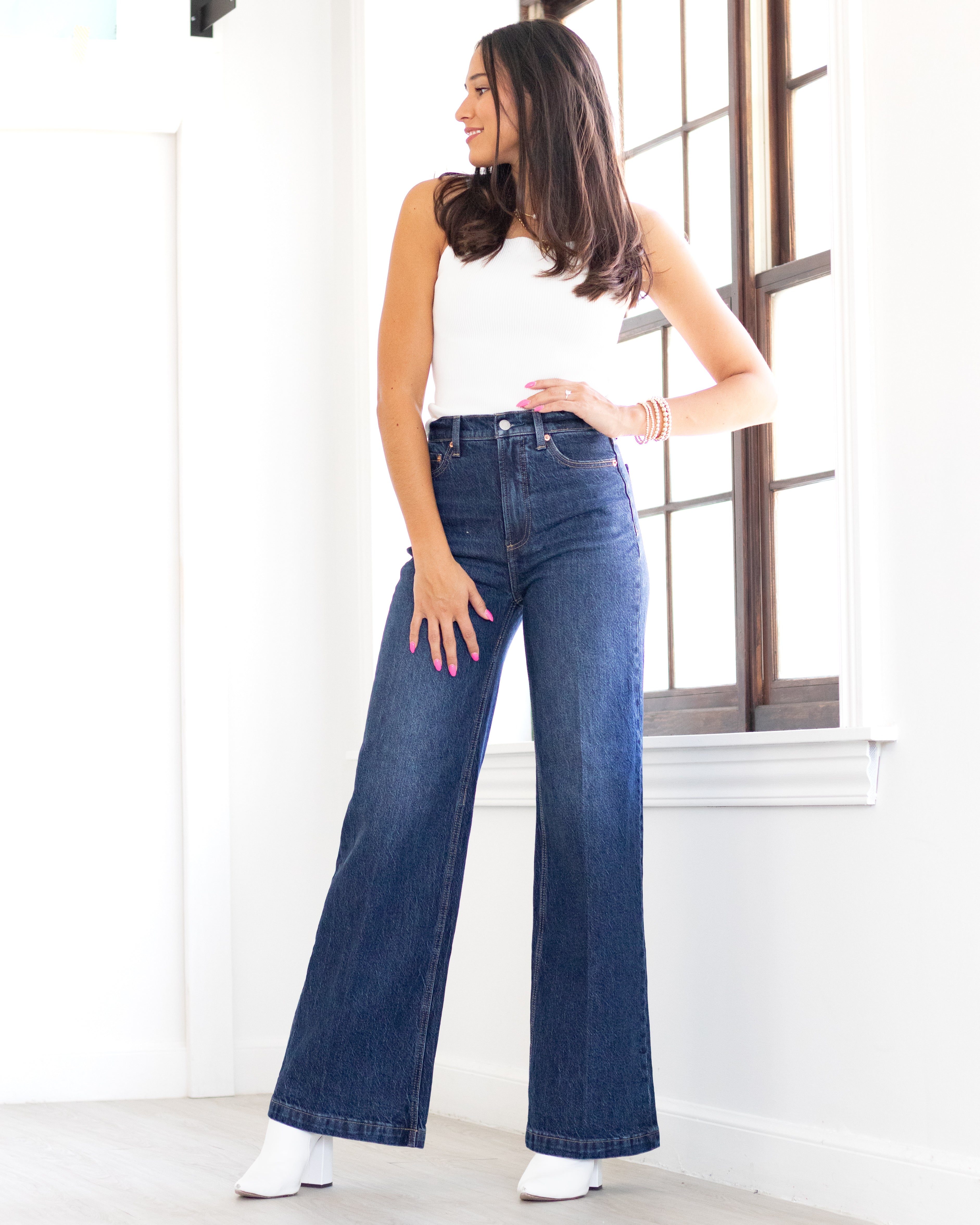 A person standing in a room with light coming through the window, wearing high-waisted, ultra-wide leg jeans and a white top.