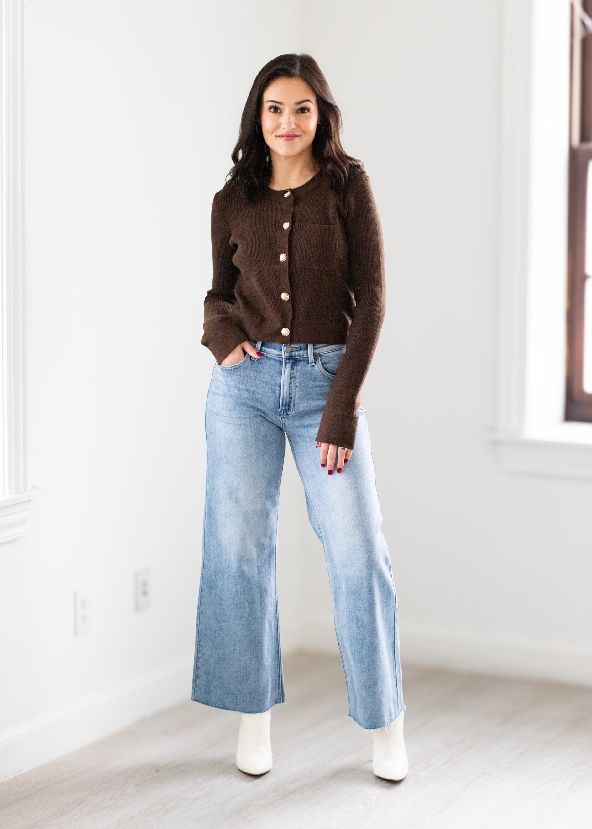 Woman wearing a brown cardigan, light blue jeans, and white boots in a minimalistic room.
