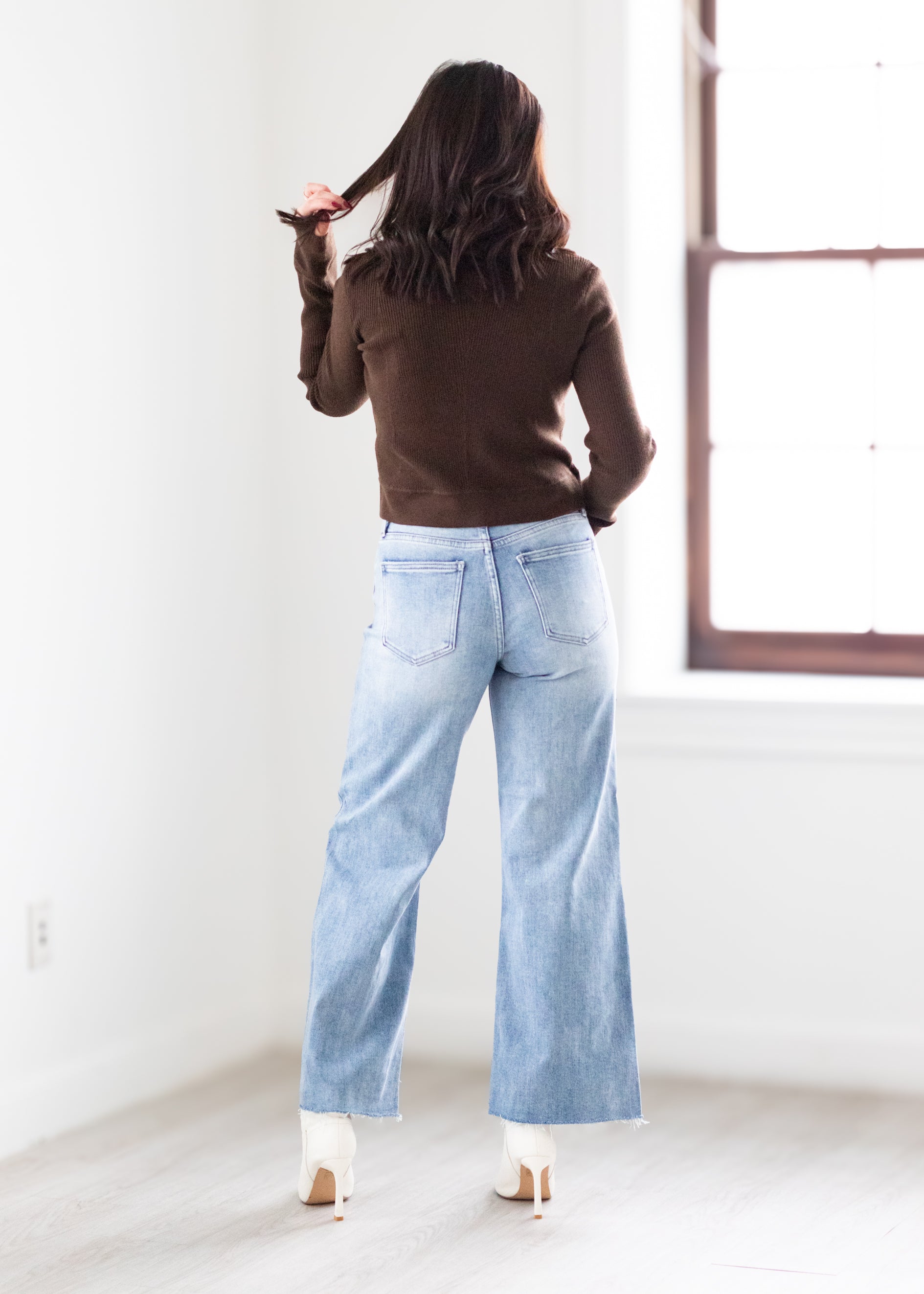 Person wearing a brown sweater and light blue jeans standing in a room with a window.