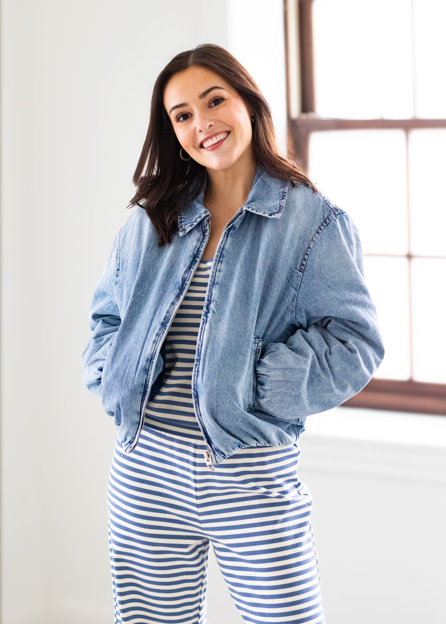 Woman wearing a denim jacket over a striped outfit in a bright room.