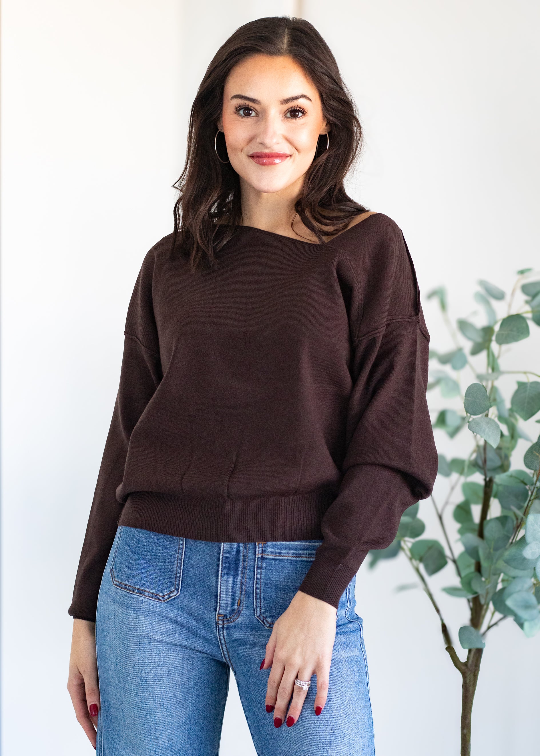 Woman wearing a brown sweater and blue jeans standing against a white background with a plant.