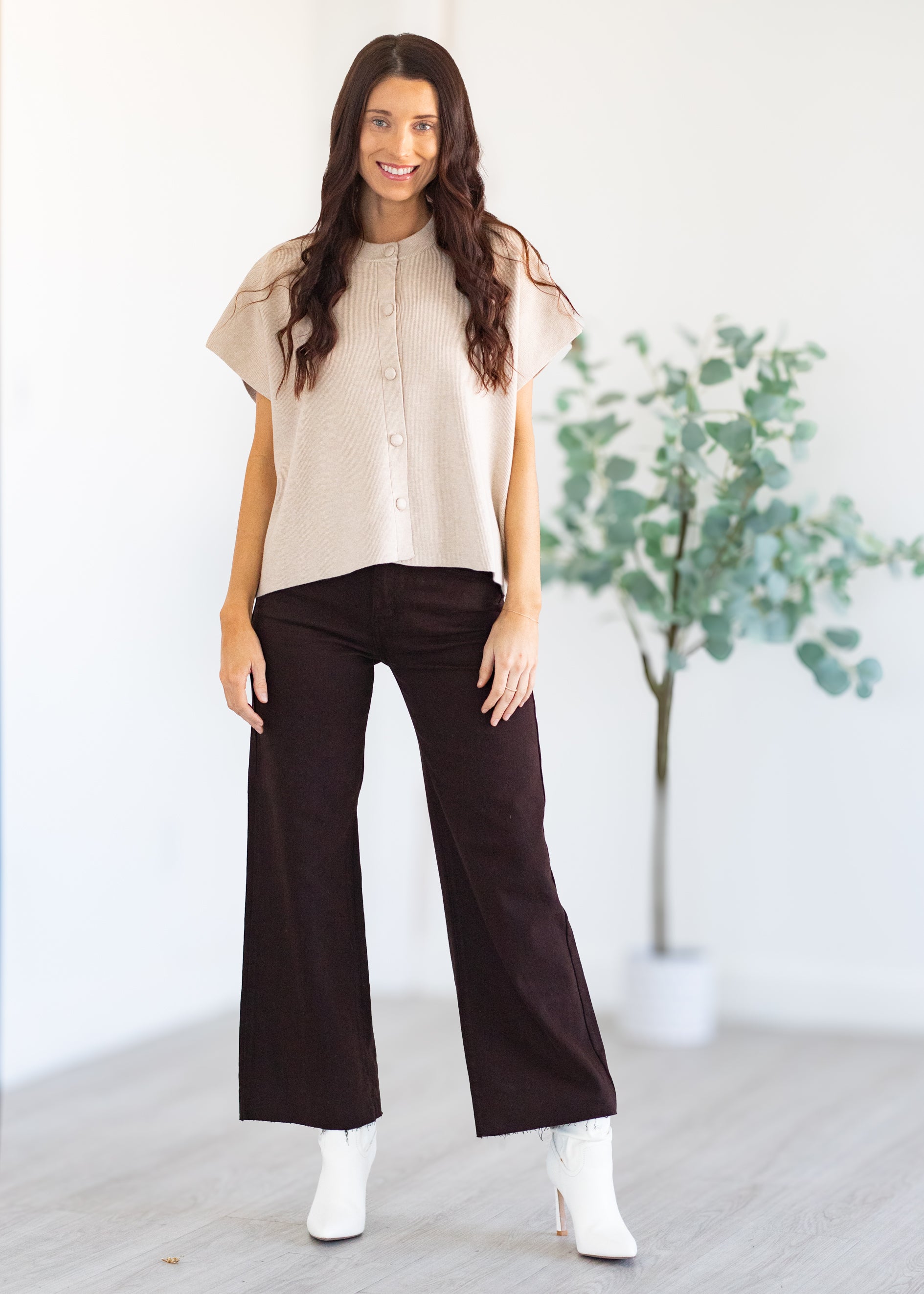 Woman wearing a beige blouse and dark pants standing in a minimalistic room with a plant.