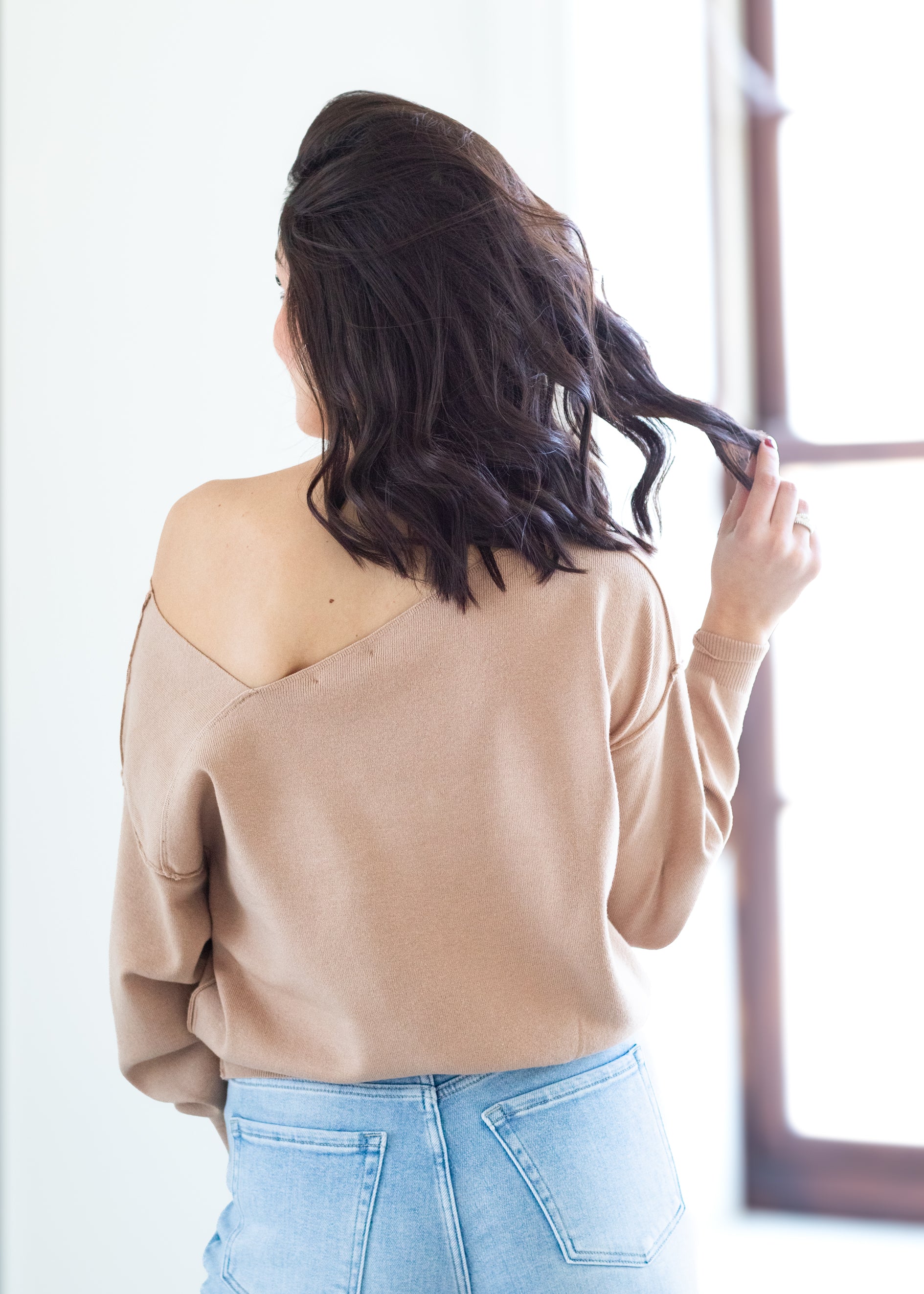 Woman wearing a beige sweater and blue jeans, standing in front of a window.