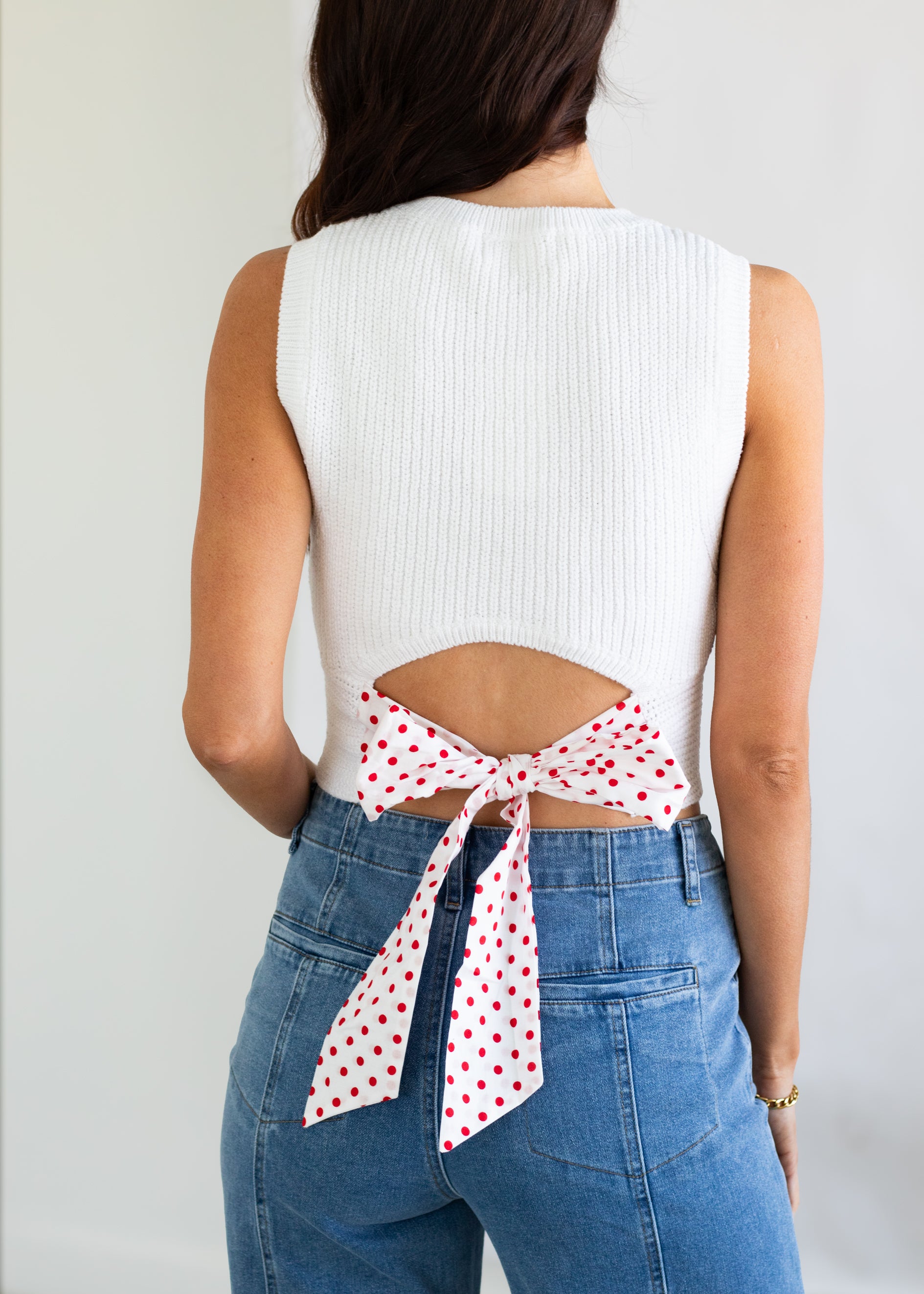 Person wearing a white sleeveless top with a red polka dot ribbon and blue jeans on a plain background