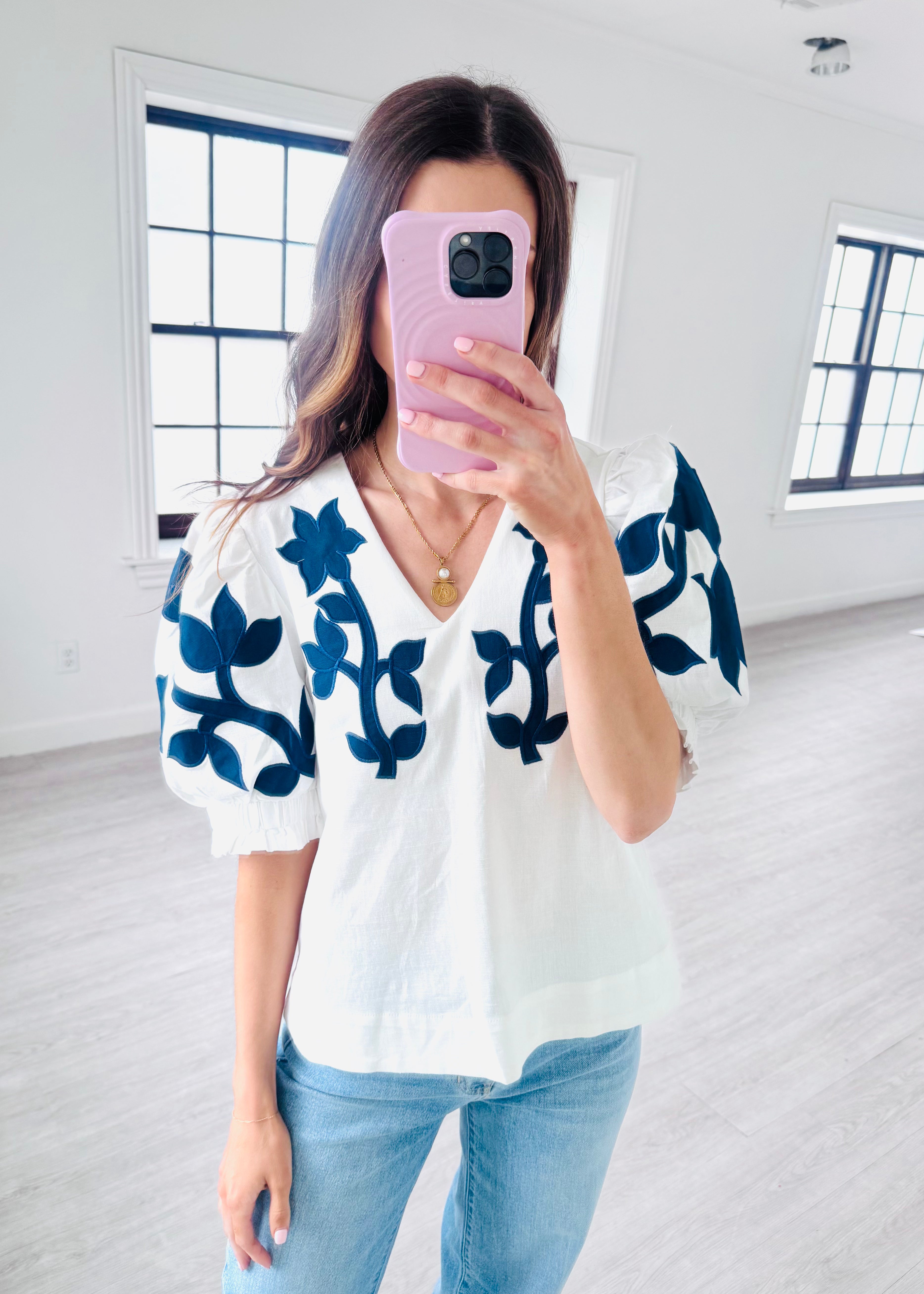 Woman taking a mirror selfie wearing a white blouse with blue patterns and light blue jeans.