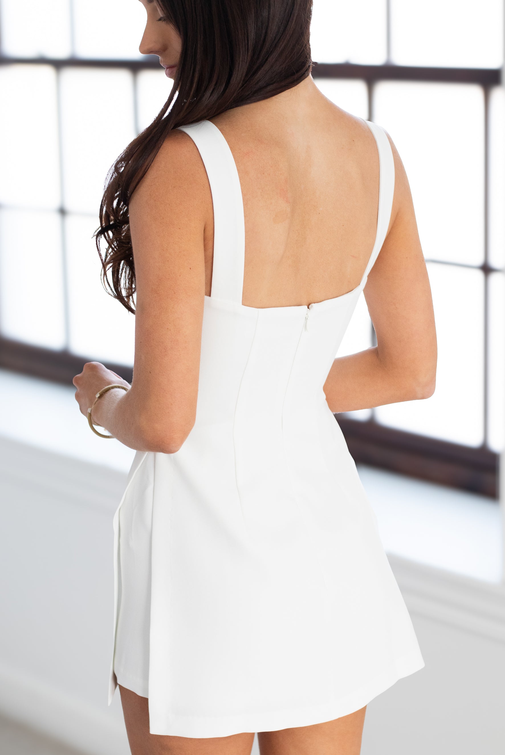Woman wearing a white dress with a backless design, standing in front of a window.