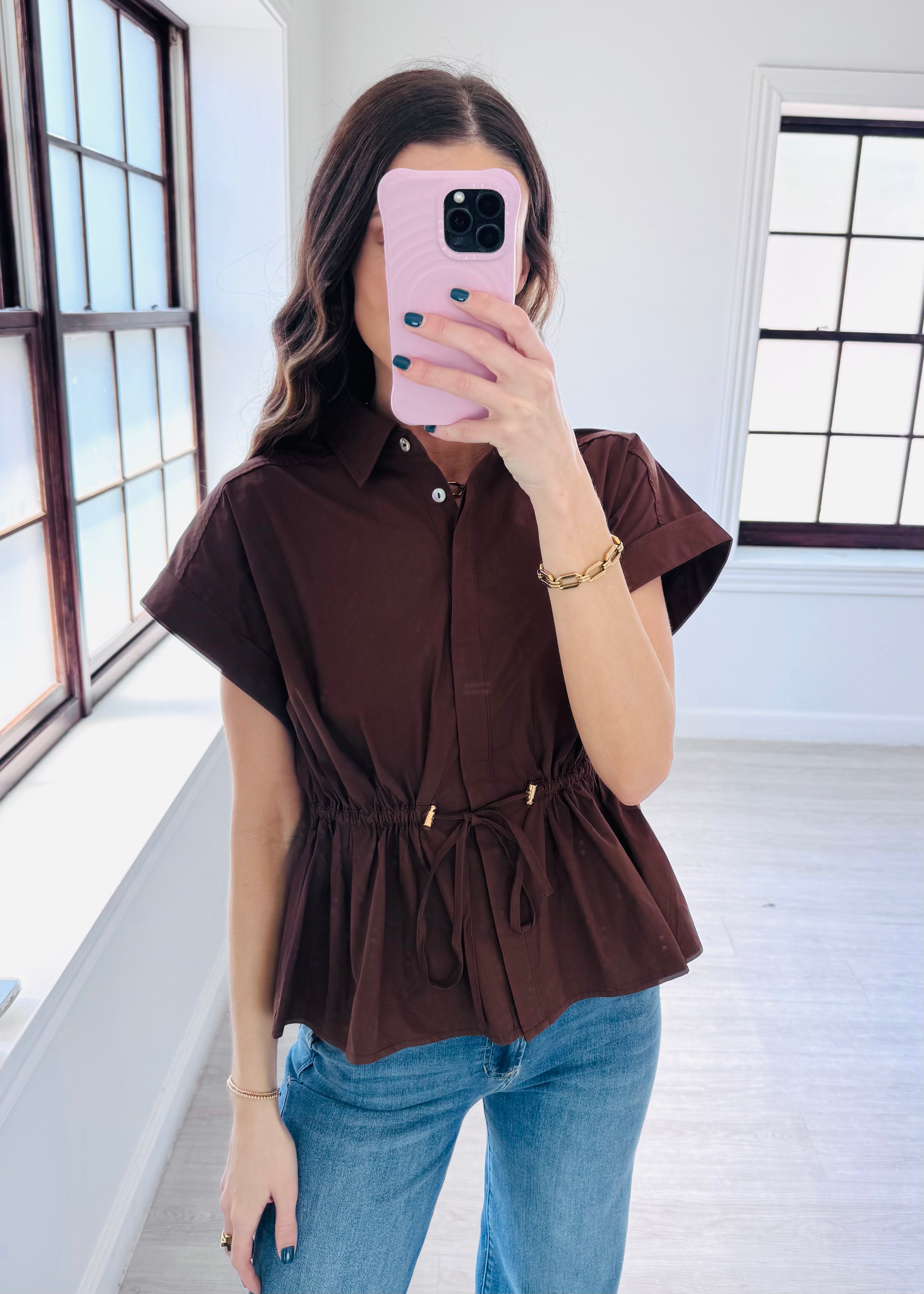A woman takes a mirror selfie wearing a dark brown short-sleeve button-up blouse with a drawstring waist and blue jeans in a bright room with large windows.