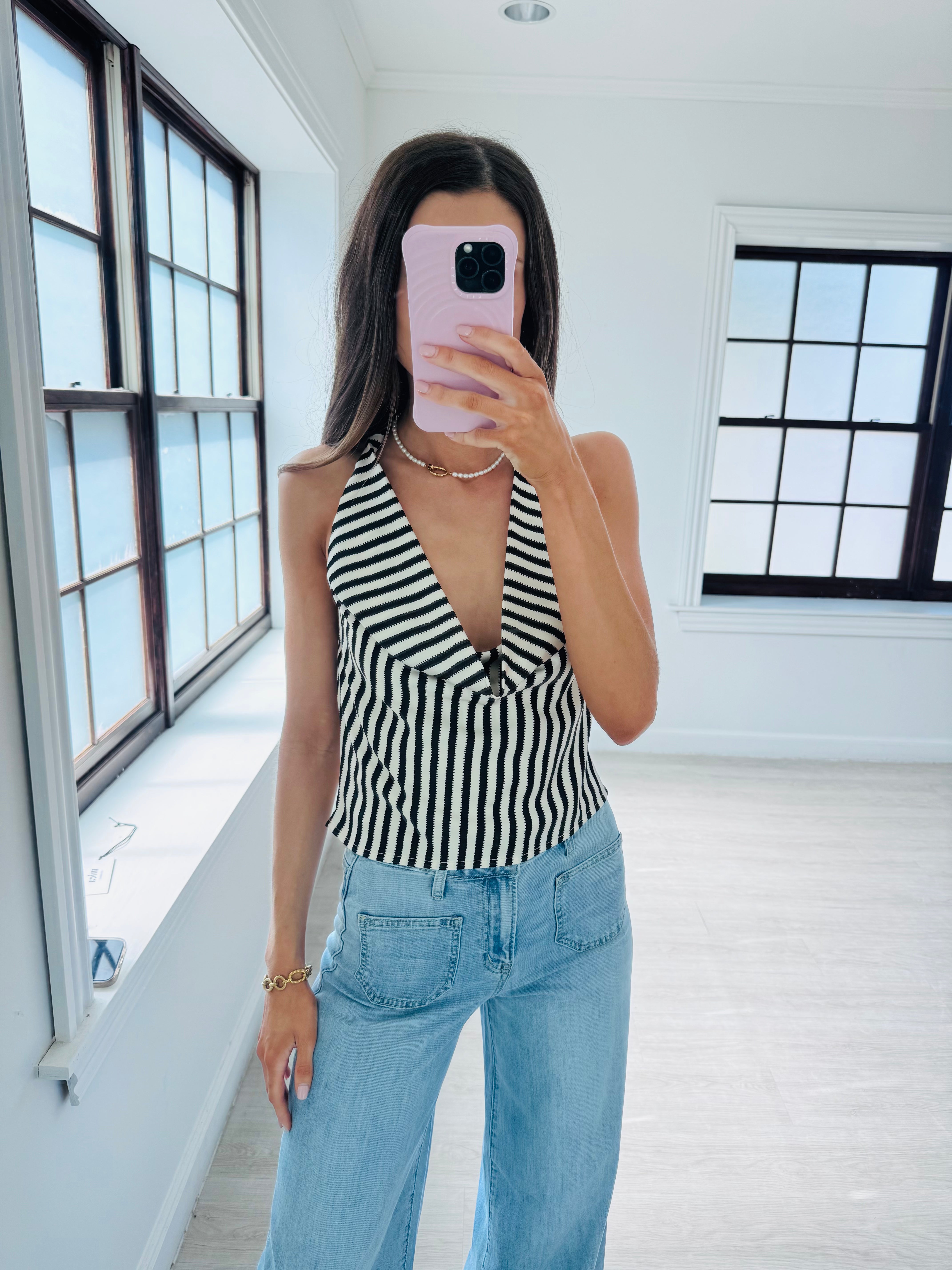 A woman takes a mirror selfie wearing a black and white striped halter top and light blue jeans in a bright, modern room with large black-framed windows.