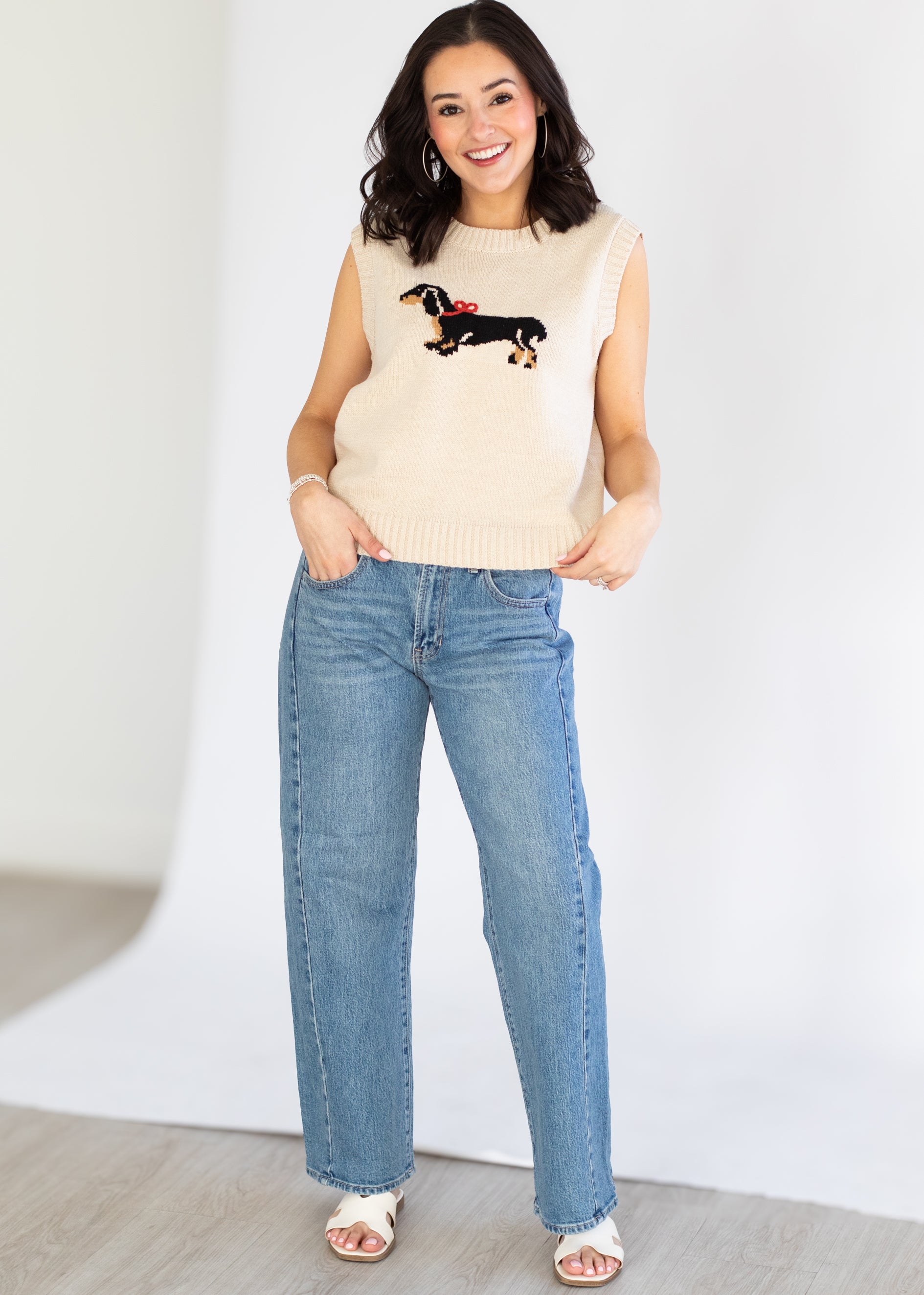 Woman wearing a beige sleeveless sweater with a dog design and blue jeans against a white background