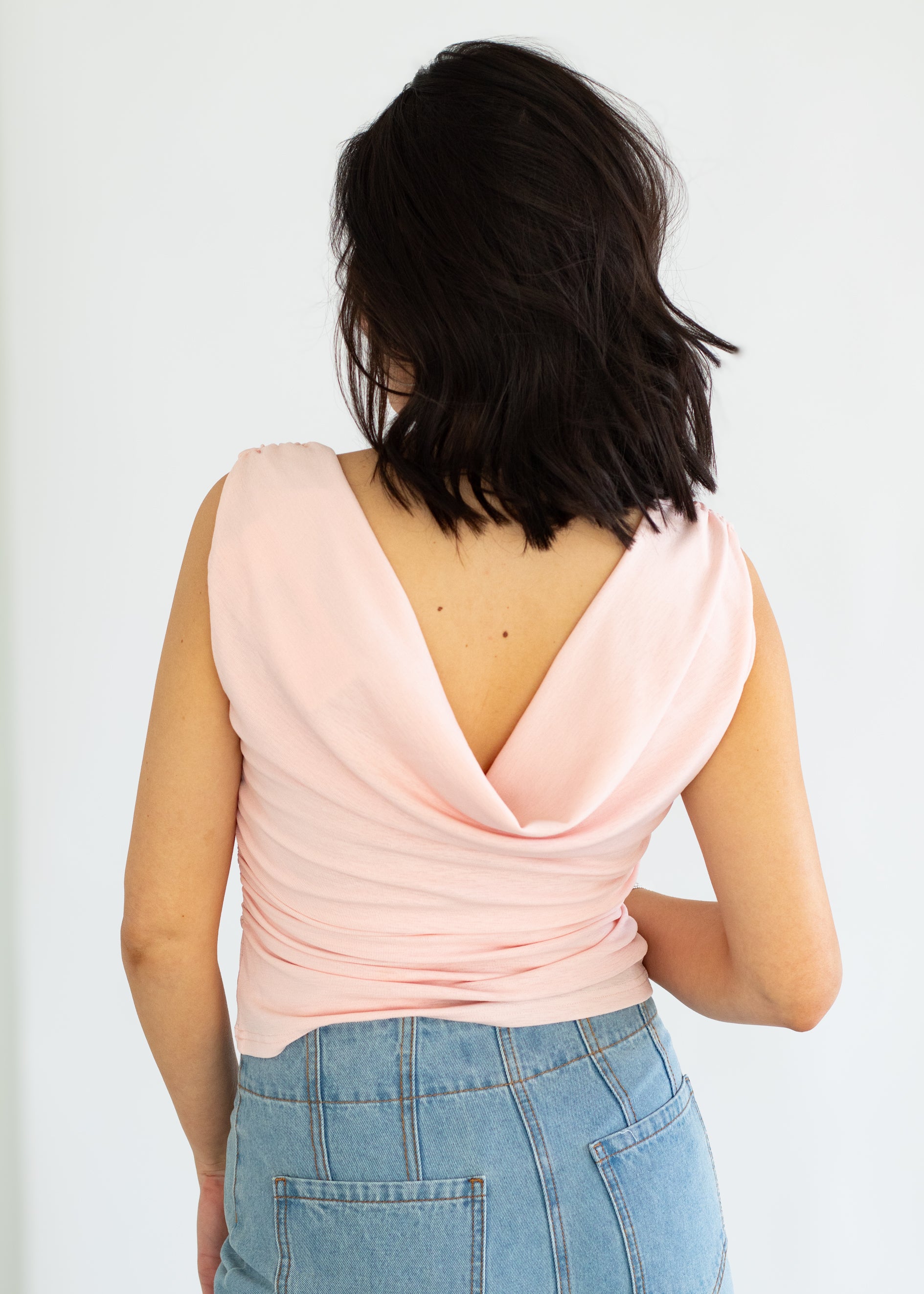 Person wearing a light pink sleeveless top with a V-neckline and blue jeans on a white background