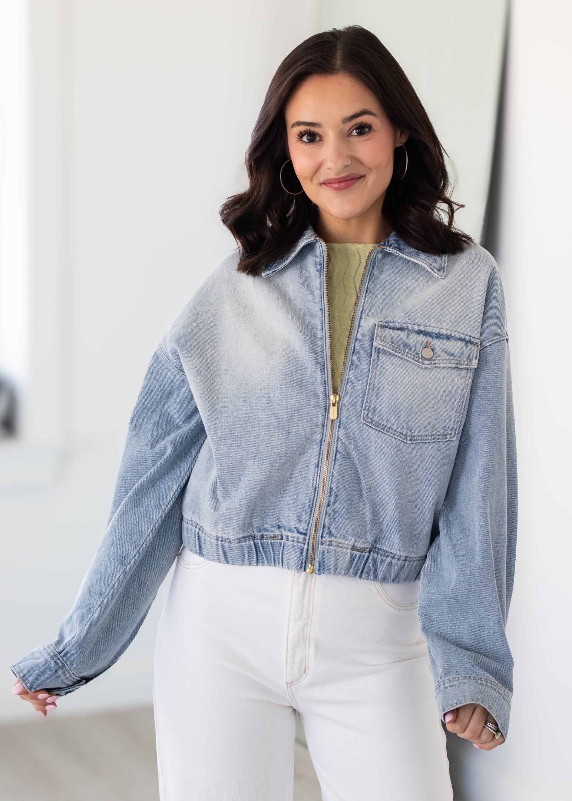 Woman wearing a light blue denim jacket over a green top and white pants, standing against a plain background.