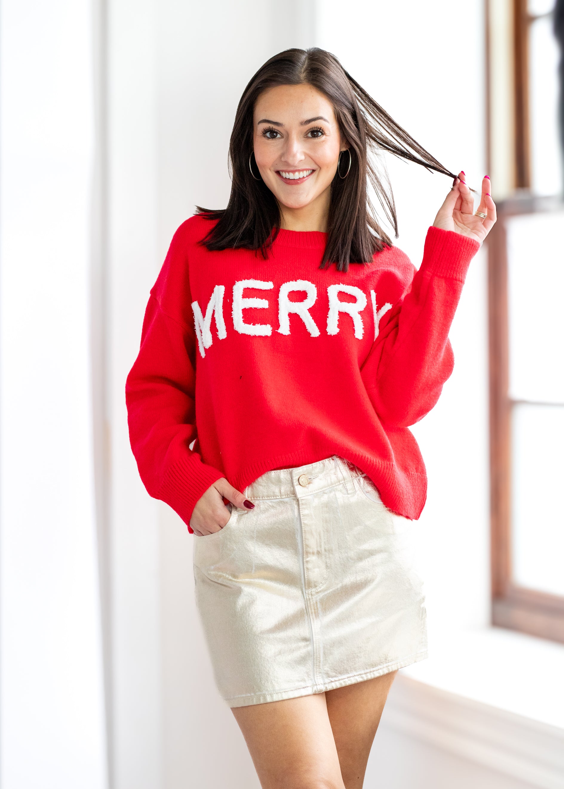 Woman wearing a red 'MERRY' sweater and gold skirt indoors.