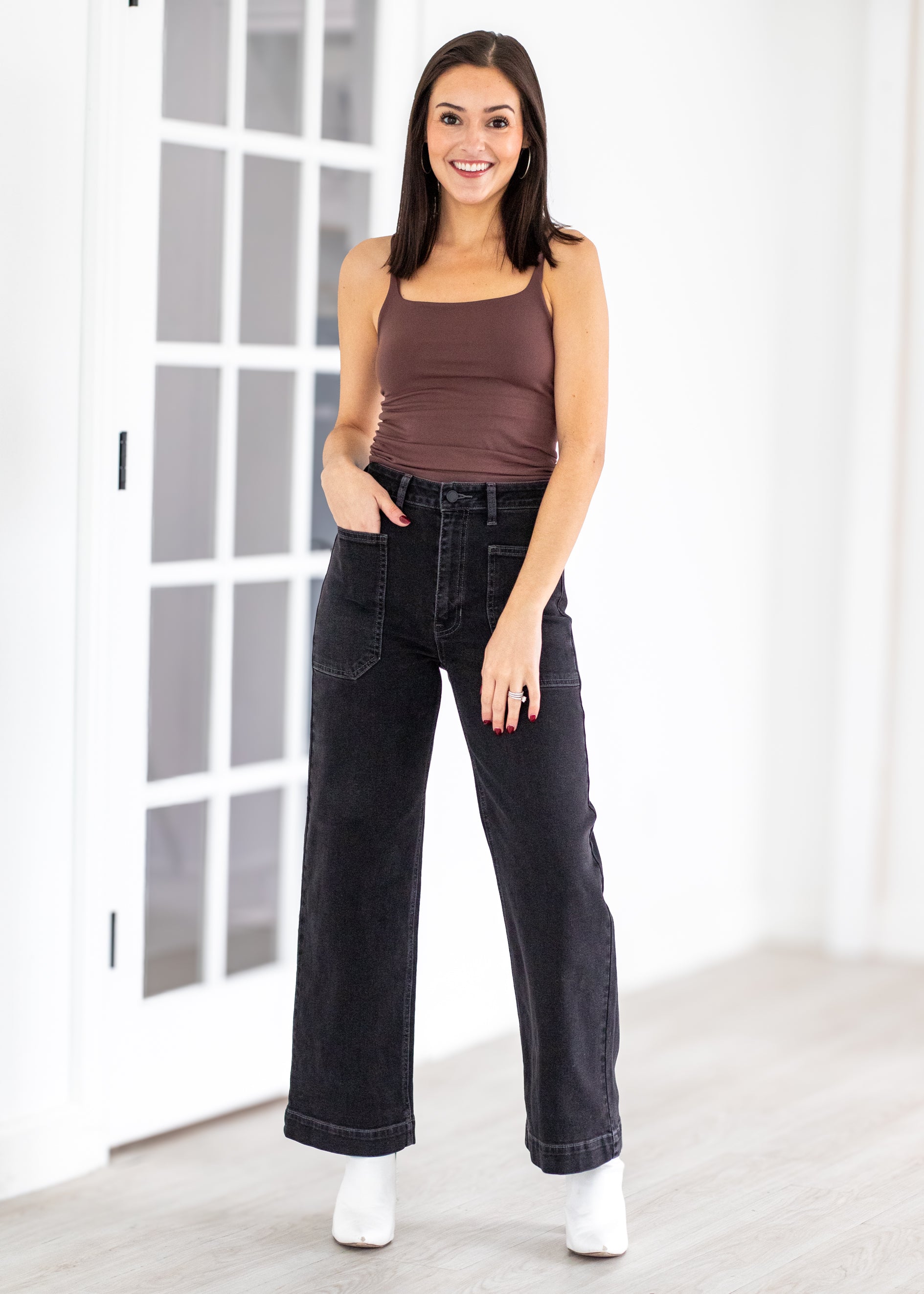 Woman wearing a brown tank top and black jeans standing in a room with white walls and a glass door.