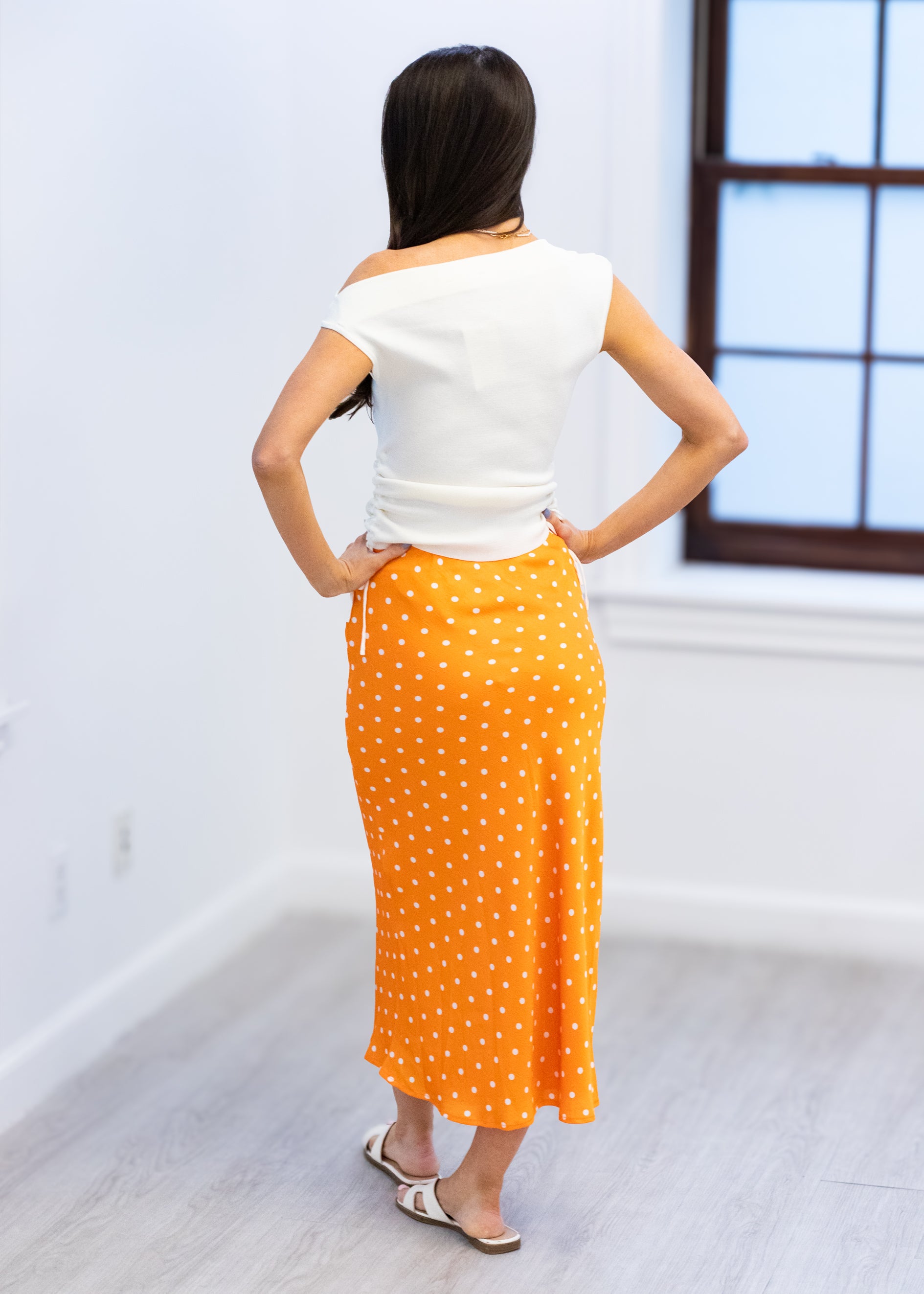 Woman wearing a white top and orange polka dot skirt in a room with a window.