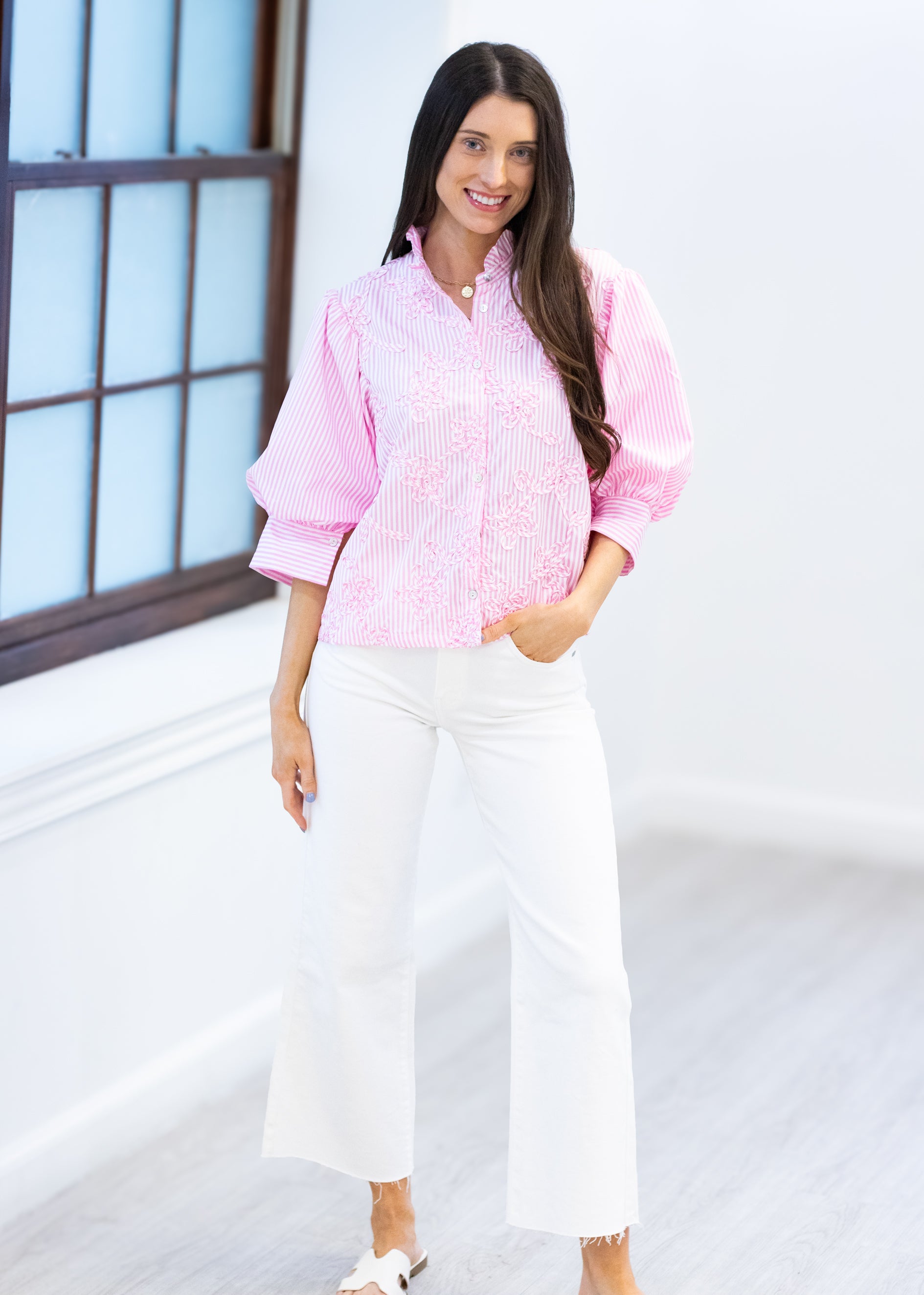 Woman wearing a pink checkered shirt and white pants standing against a light-colored wall.