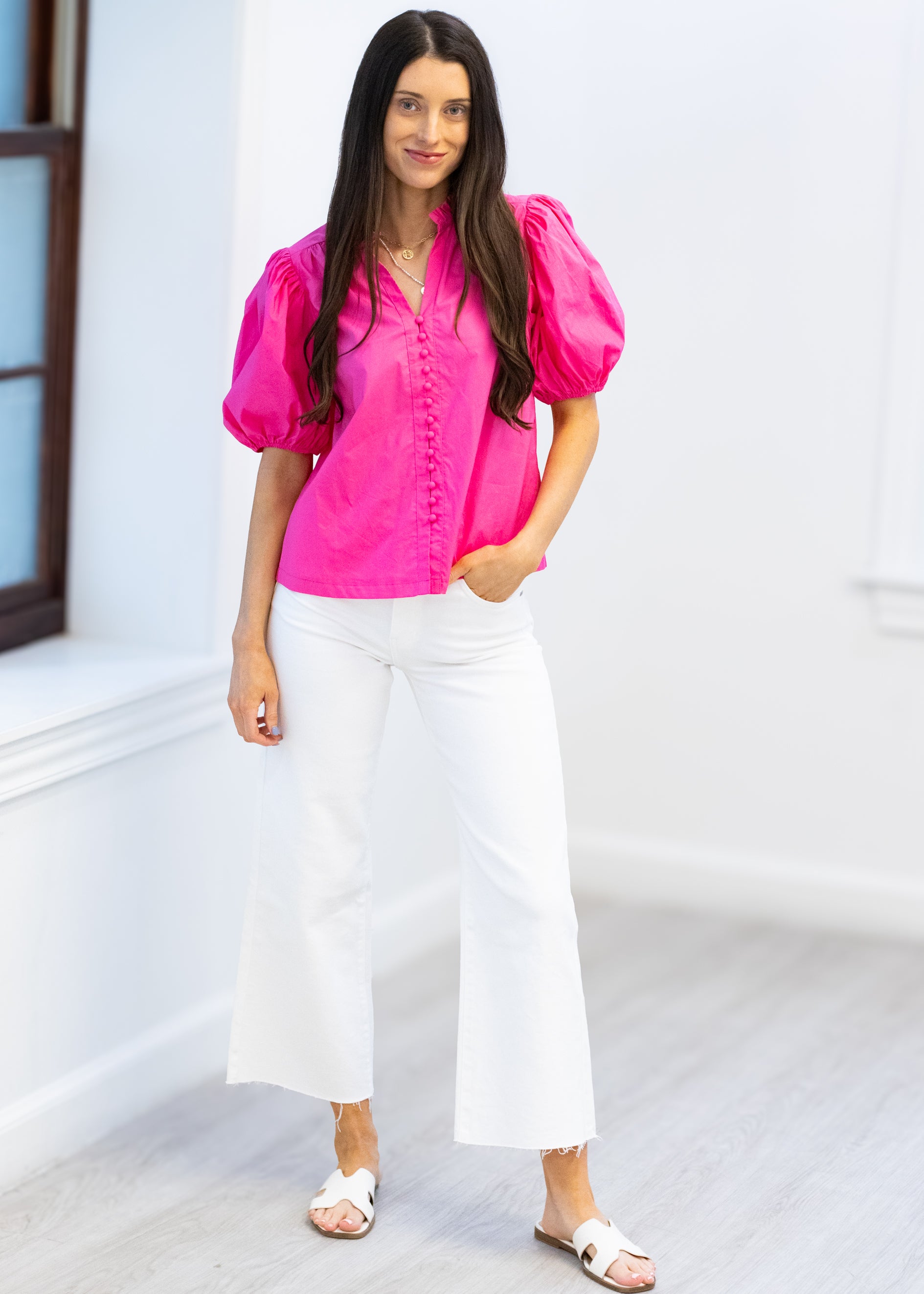 Woman wearing a bright pink blouse and white pants standing in a minimalistic room.