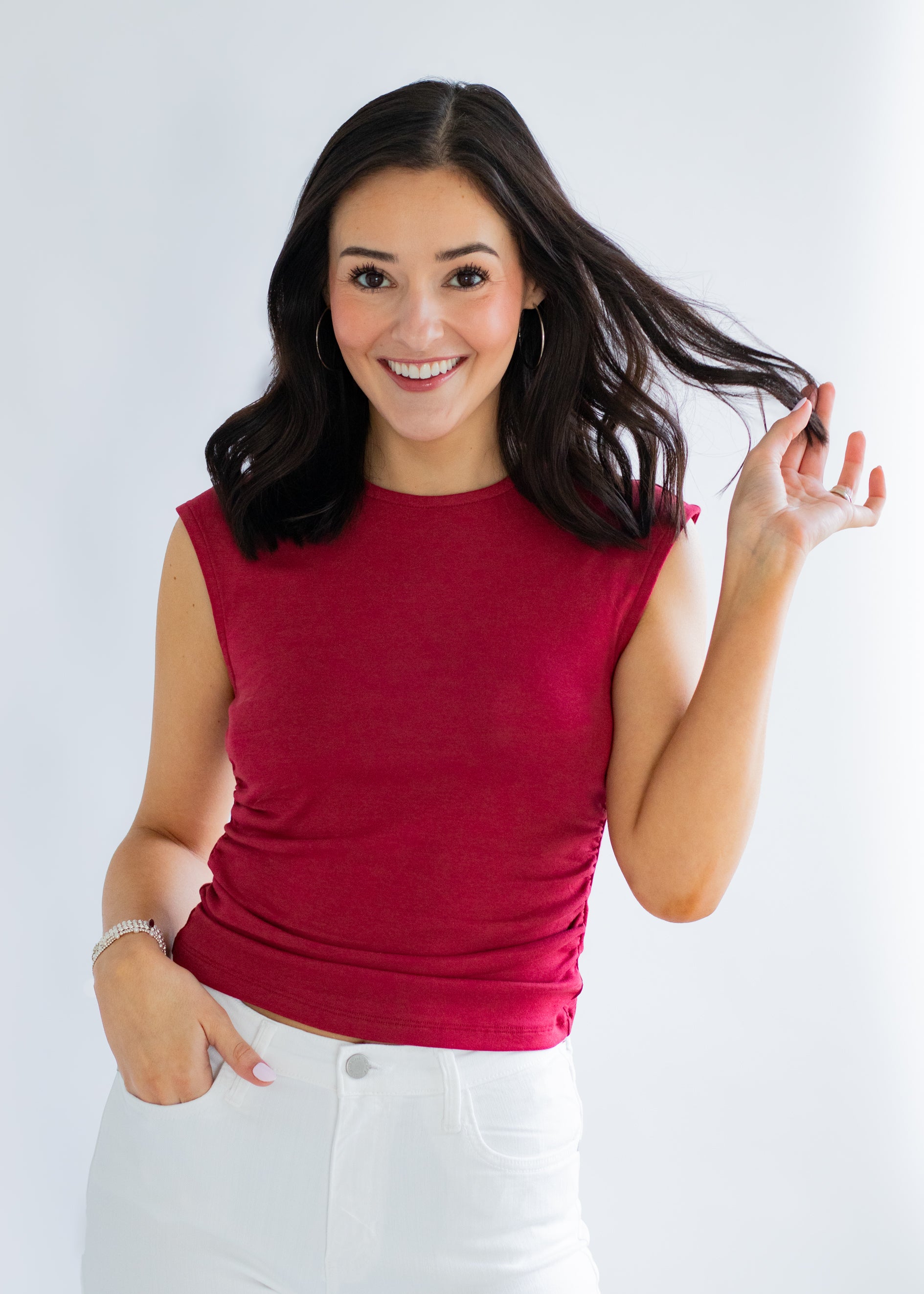 Woman wearing a red sleeveless top and white pants against a plain background