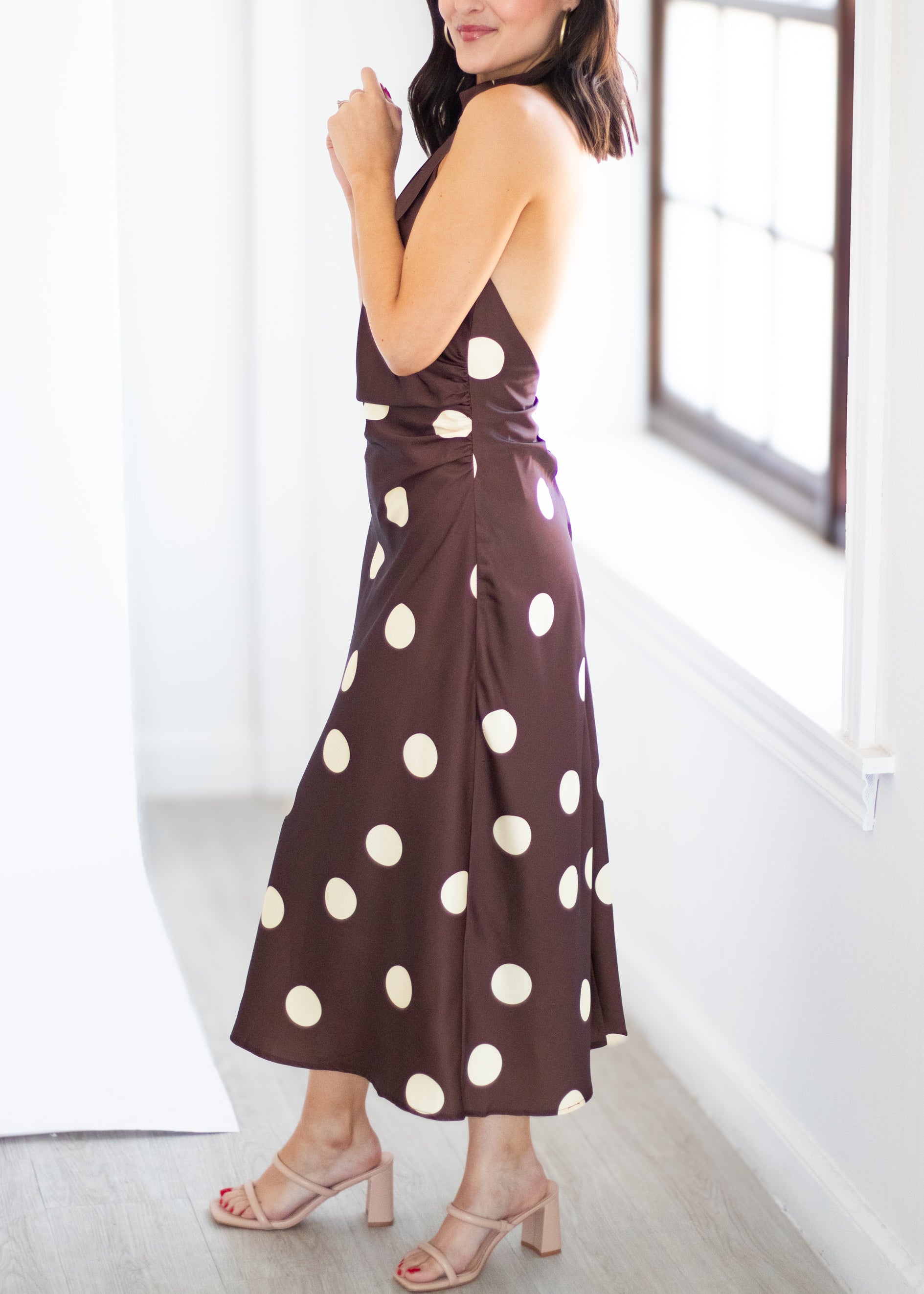 A woman wearing a brown halter midi dress with large cream polka dots, standing in a bright room with white walls and a window.