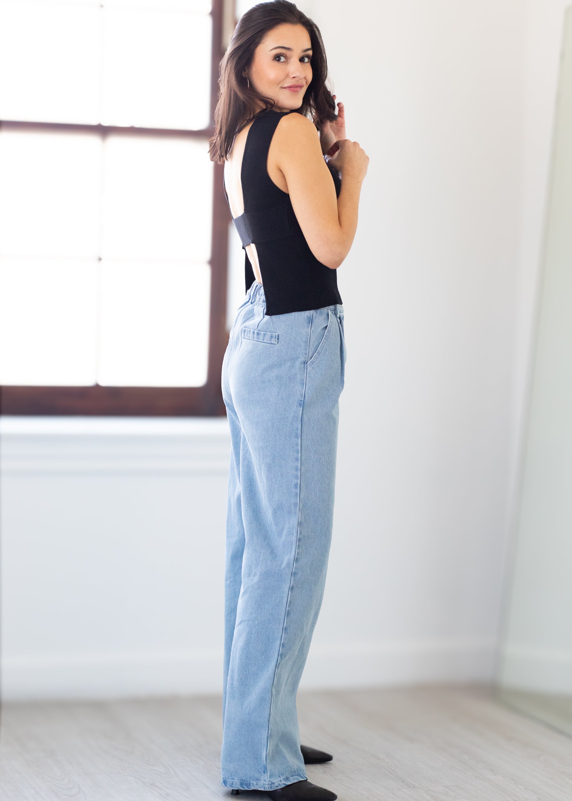 Woman wearing a black sleeveless top and light blue jeans in a room with a window.