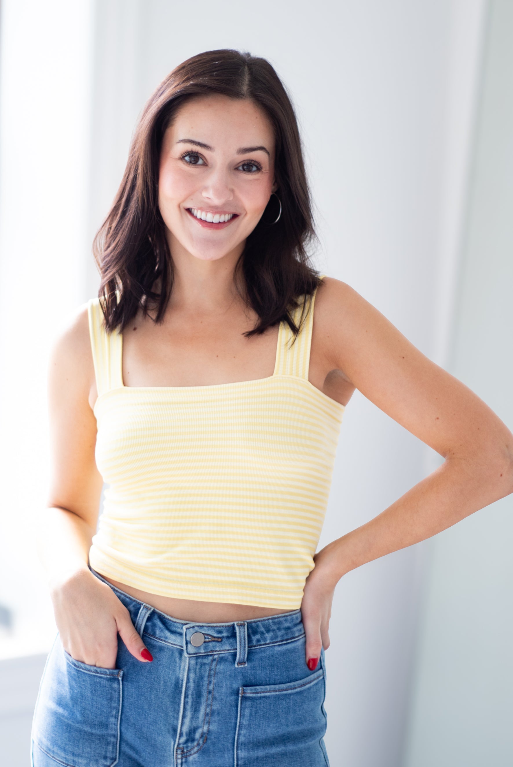 A smiling woman with dark brown hair wearing a pale yellow ribbed tank top and blue jeans, standing in a brightly lit indoor setting.