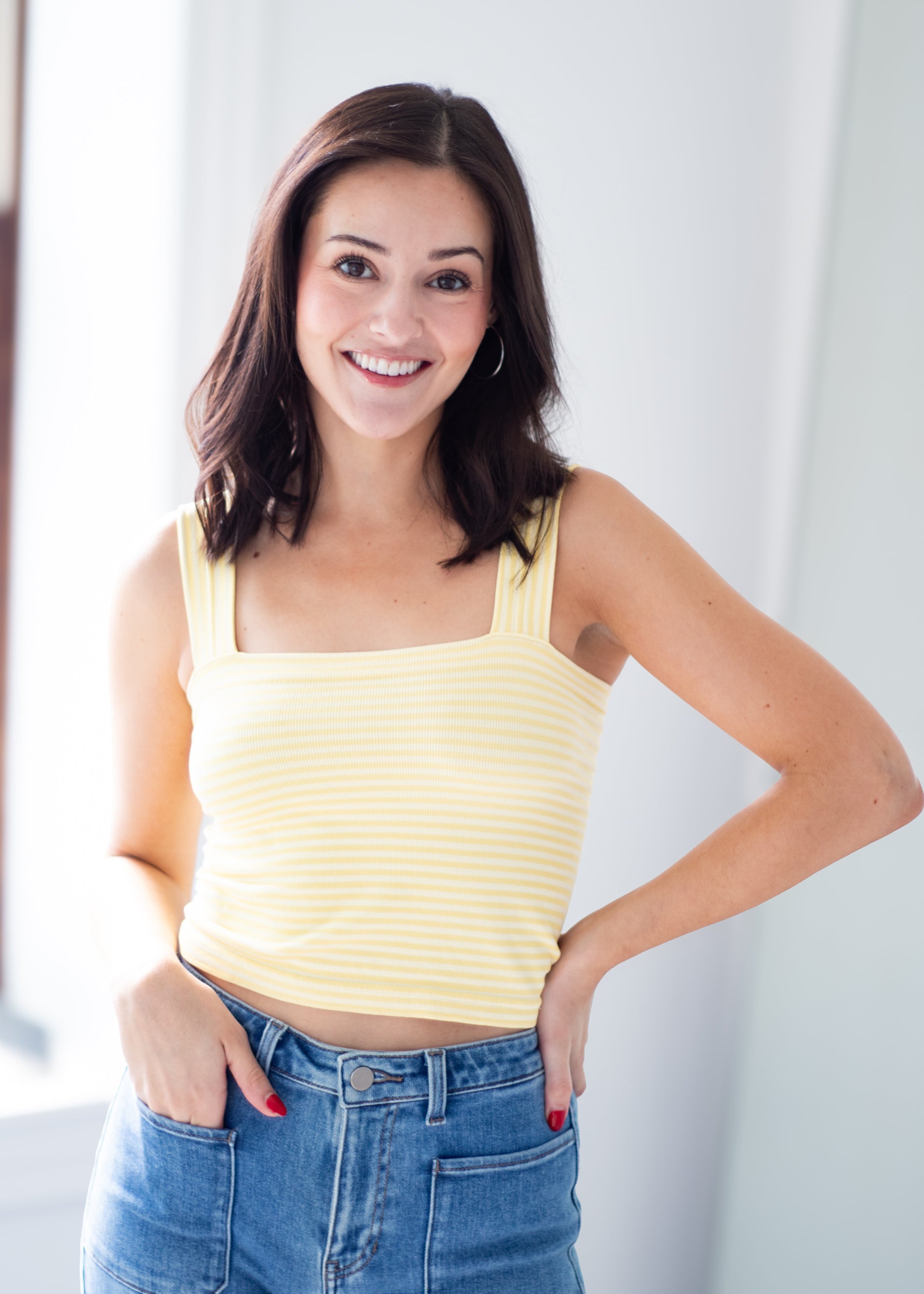 A smiling woman with dark brown hair wearing a pale yellow ribbed tank top and blue jeans, standing in a brightly lit indoor setting.