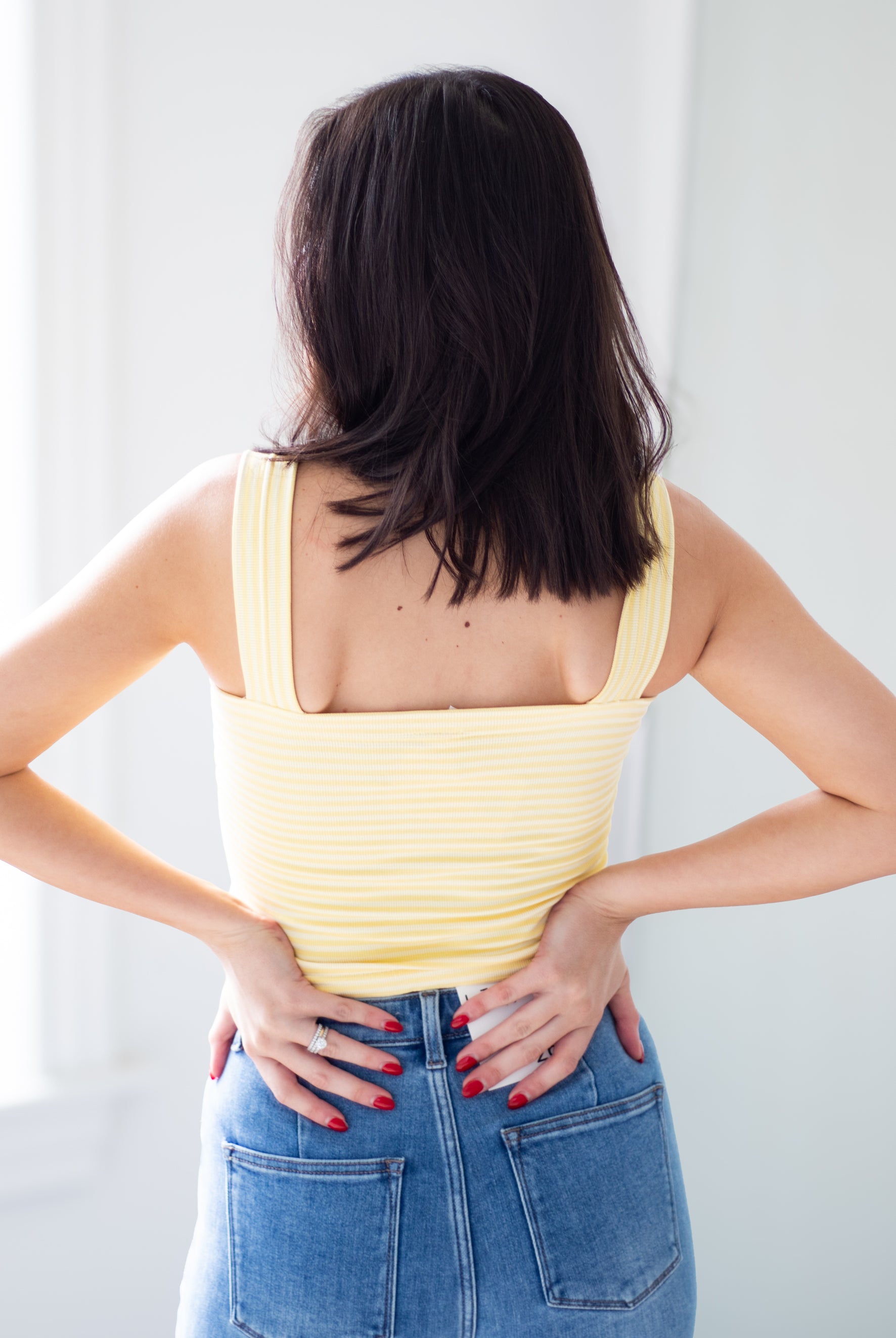 A woman viewed from behind wearing a pale yellow ribbed tank top and blue jeans, standing indoors with her hands on her lower back.