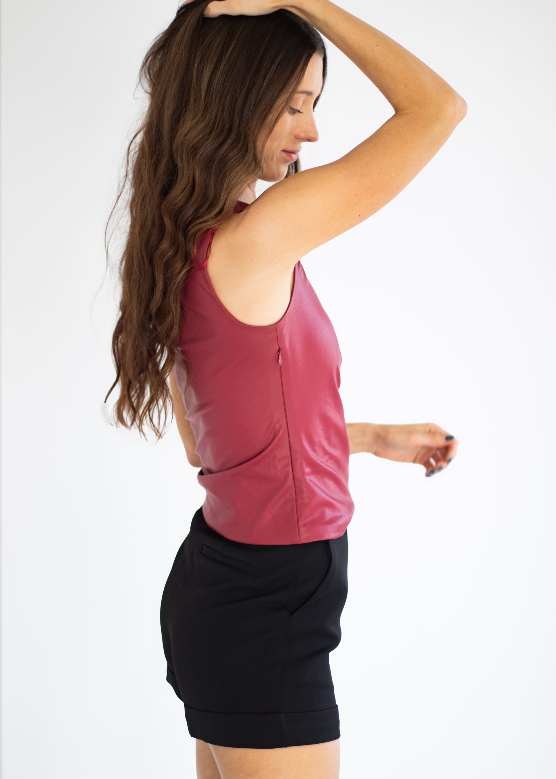 A woman wearing a sleeveless, zippered dusty rose leather-like top and black shorts, shown from a side-back angle against a plain white studio background.
