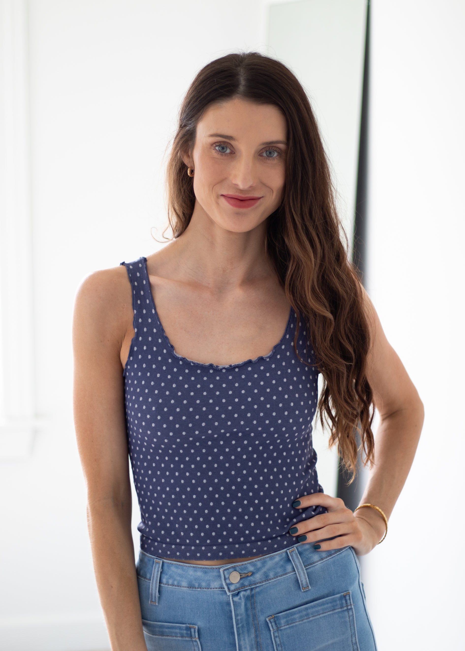 A woman with long brown hair wearing a navy blue scoop-neck tank top with small white polka dots and light blue jeans, standing in a brightly lit indoor setting.