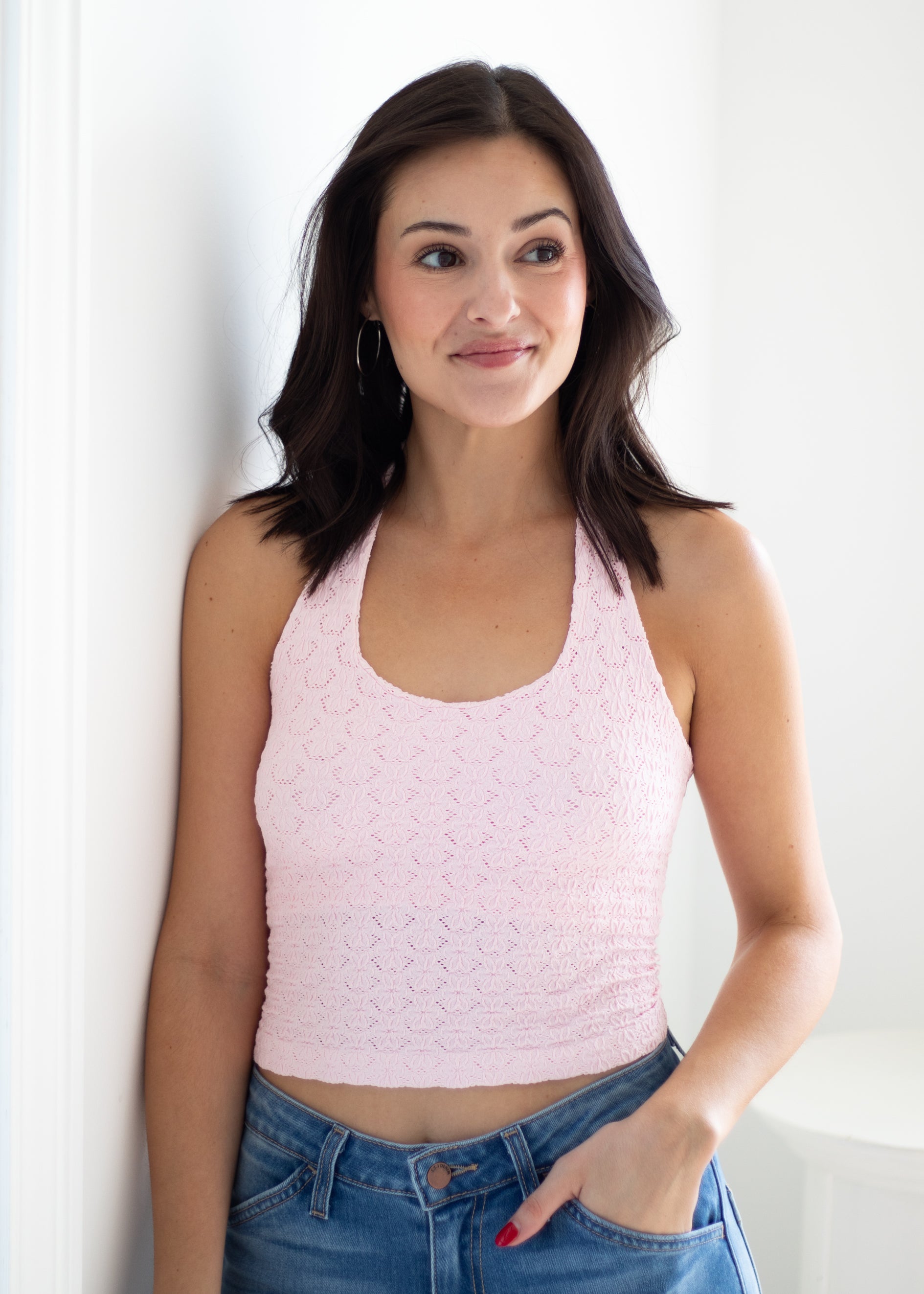A woman with dark hair wearing a light pink textured knit halter crop top and blue jeans, standing against a white wall.