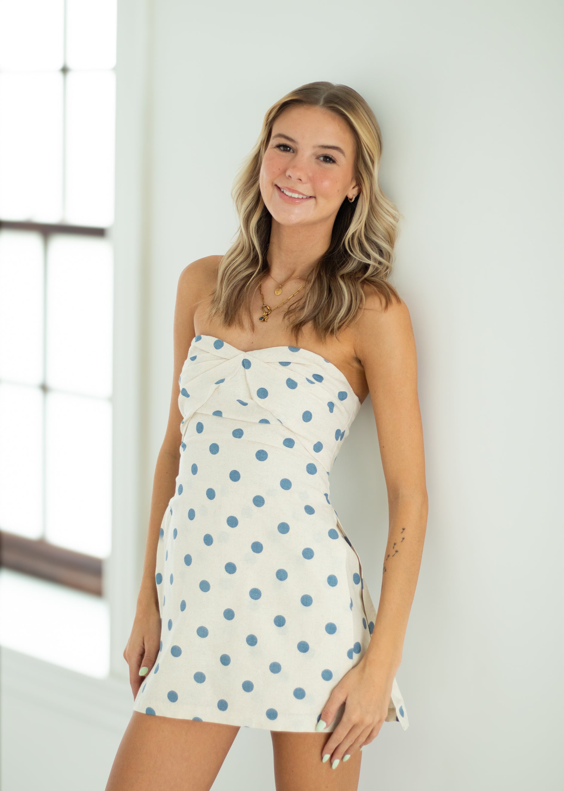Woman wearing a strapless polka dot dress against a white wall.