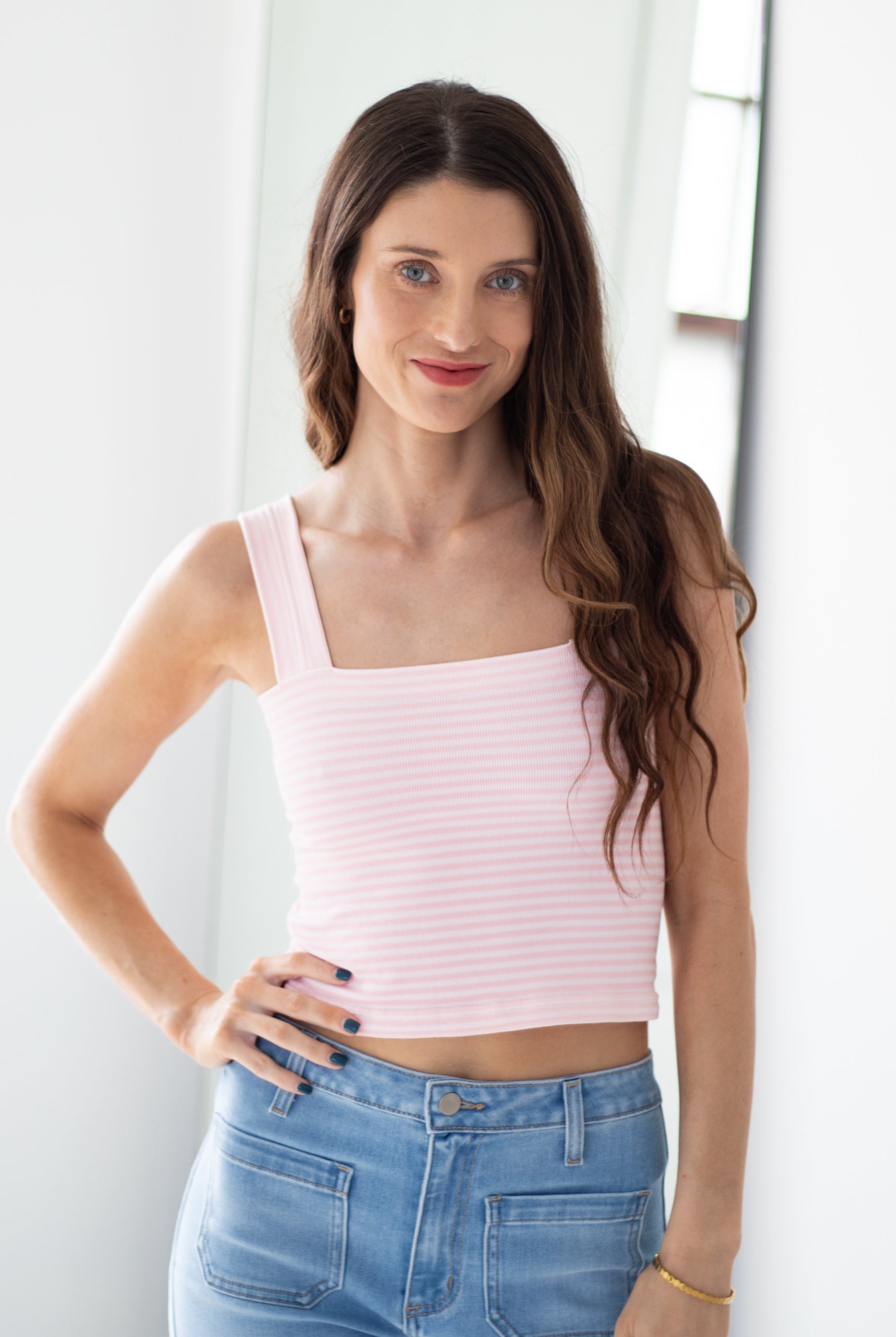 A woman with long brown hair wearing a light pink and white horizontally striped ribbed tank top and light blue jeans, posing in a brightly lit indoor setting.