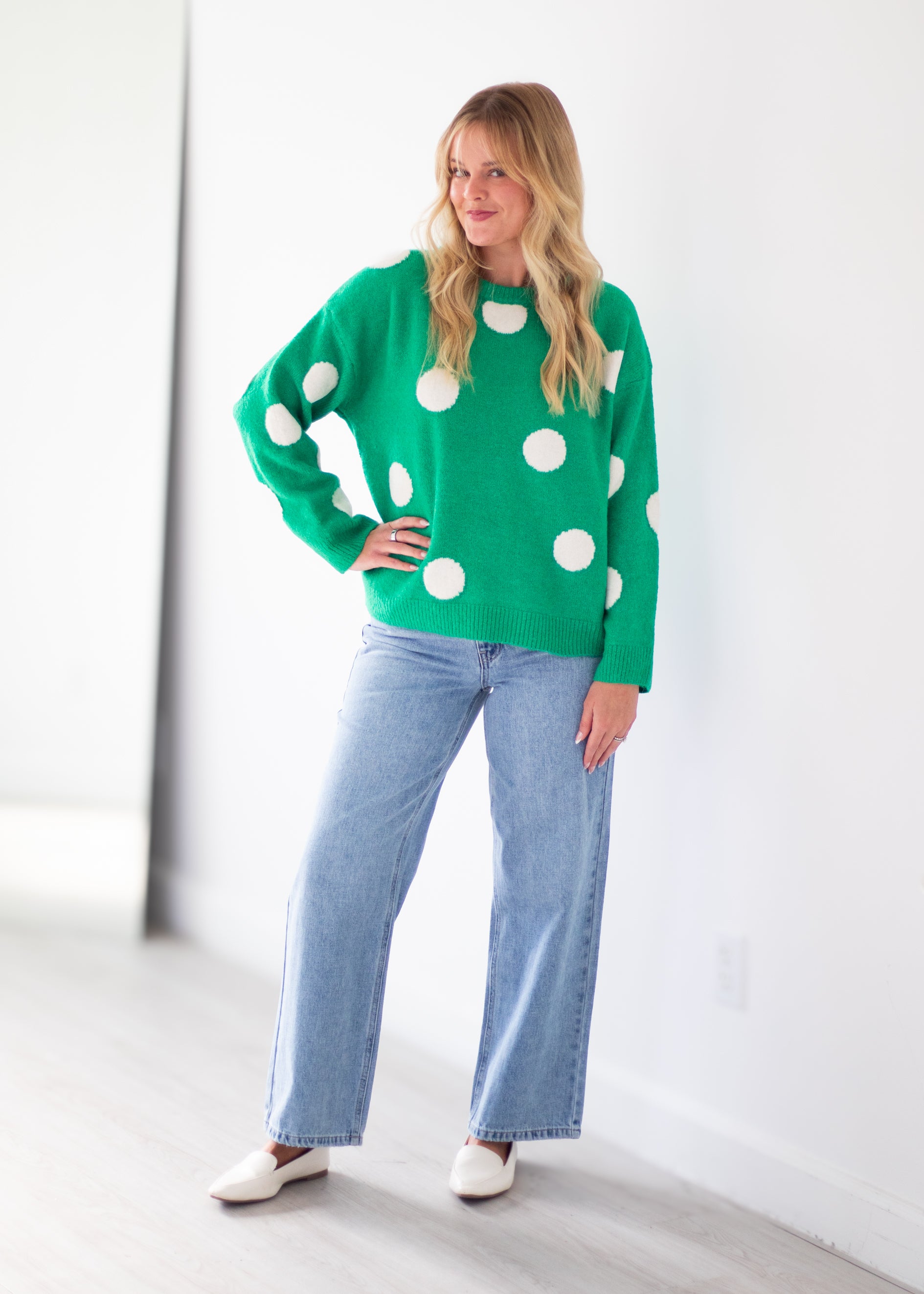 A woman modeling a bright green crewneck sweater with large white polka dots, paired with light-wash wide-leg jeans and white flats, standing in a bright, minimalist room.