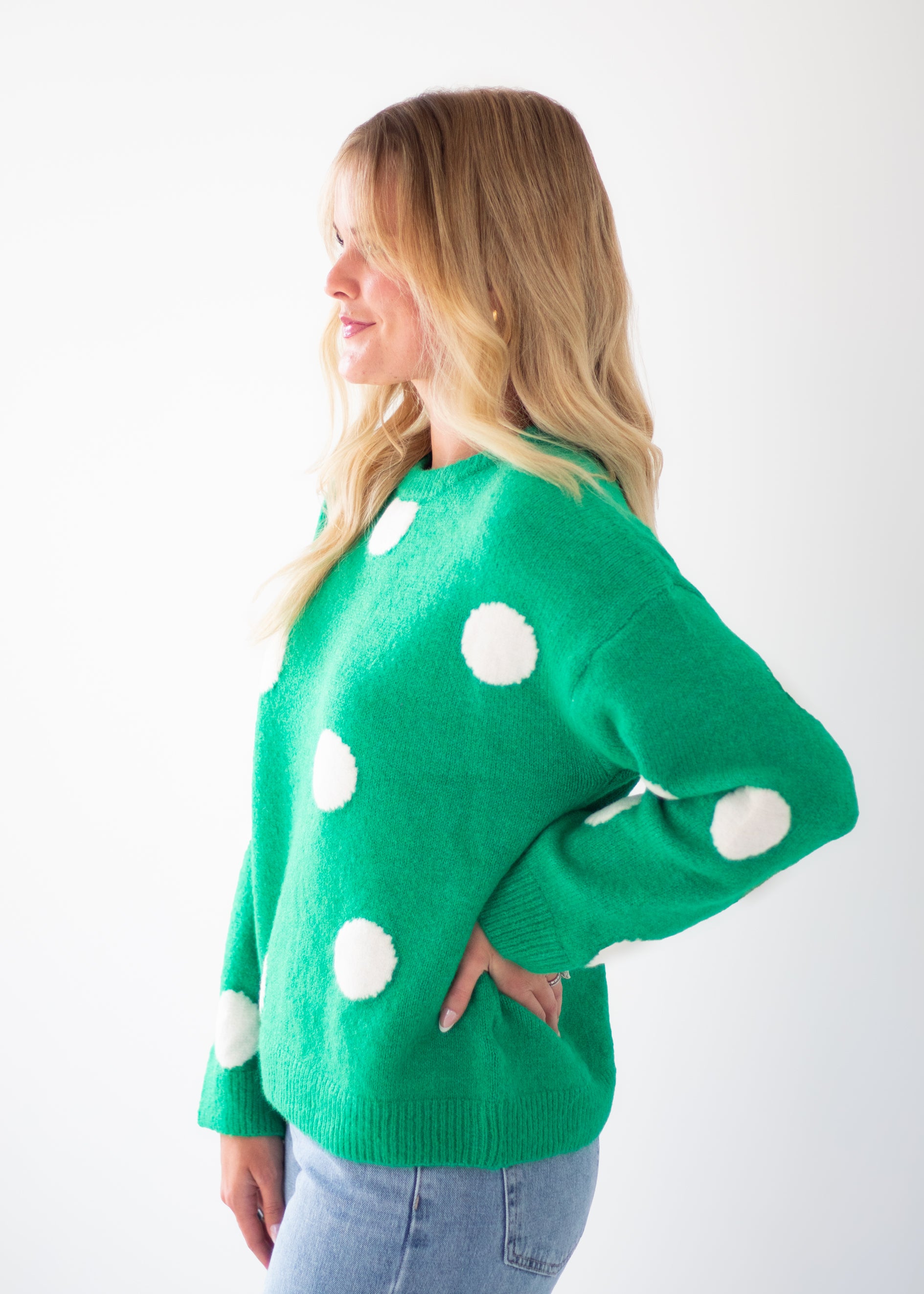 A woman wearing a bright green knit sweater with large white polka dots, paired with light blue jeans, standing against a plain white background.