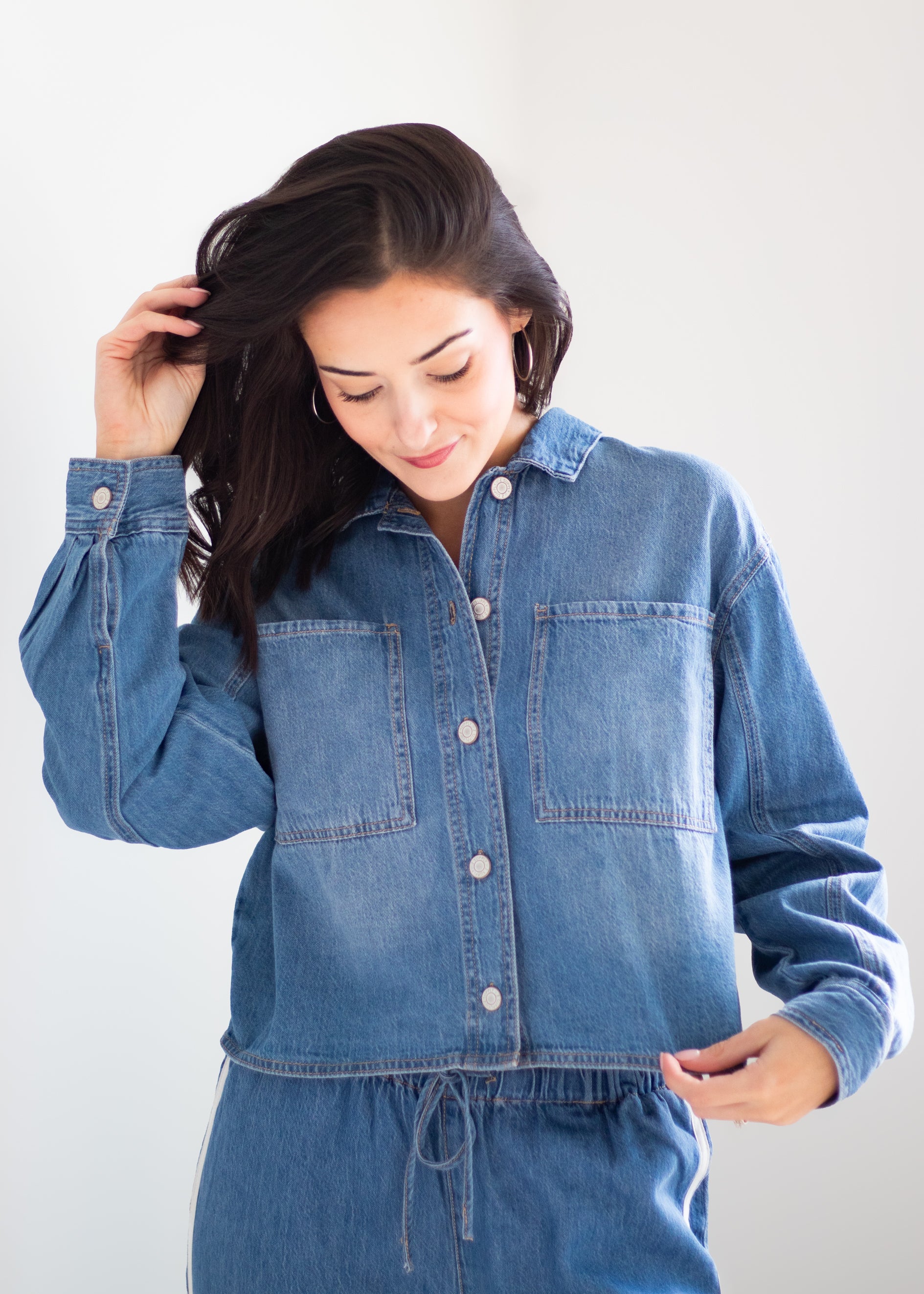A woman wearing a blue denim long-sleeve button-up shirt and matching denim drawstring pants, posing against a light gray studio background.