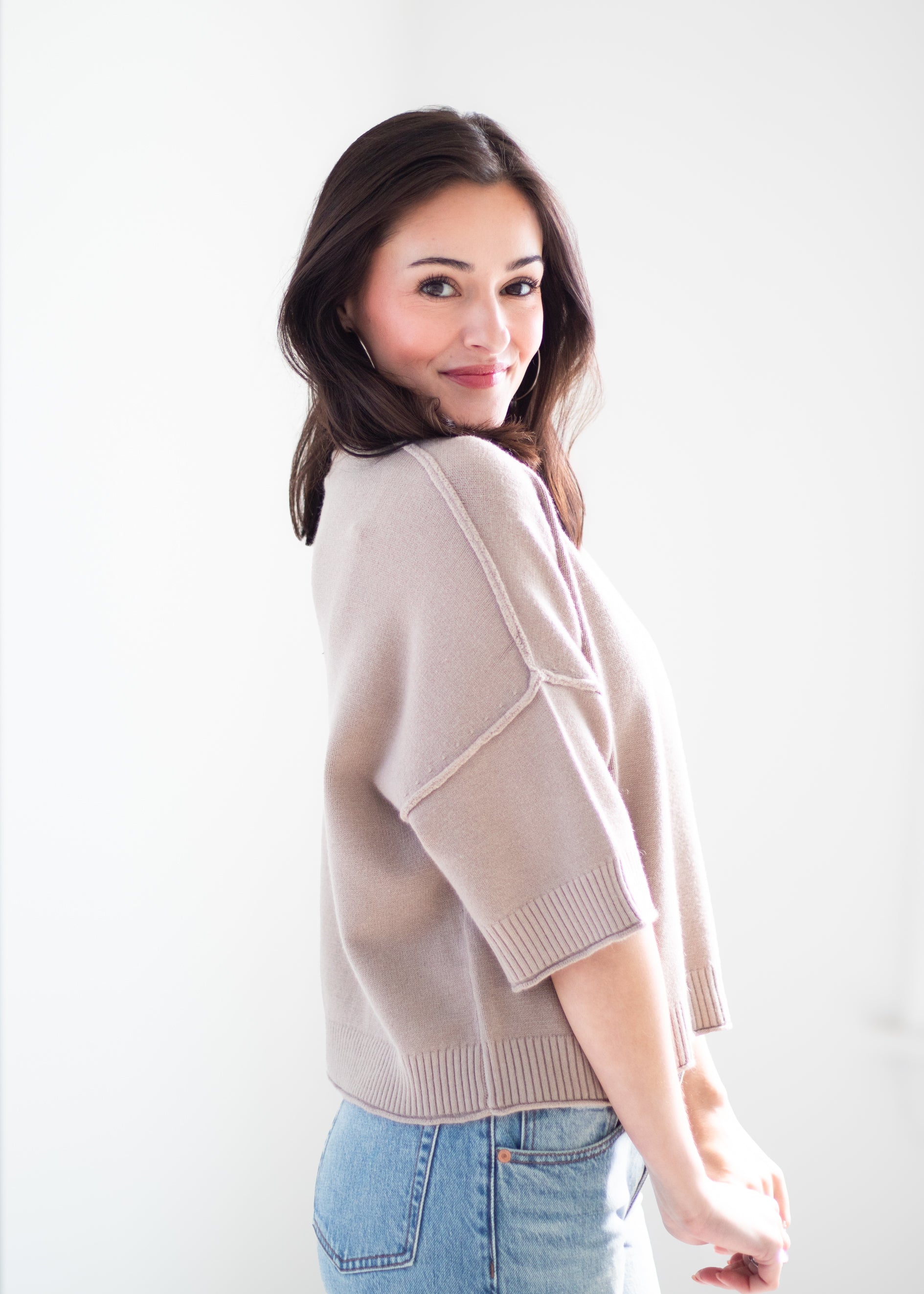 A woman wearing a light beige knit sweater with elbow-length sleeves and ribbed trim, paired with light blue jeans, photographed against a plain white background.