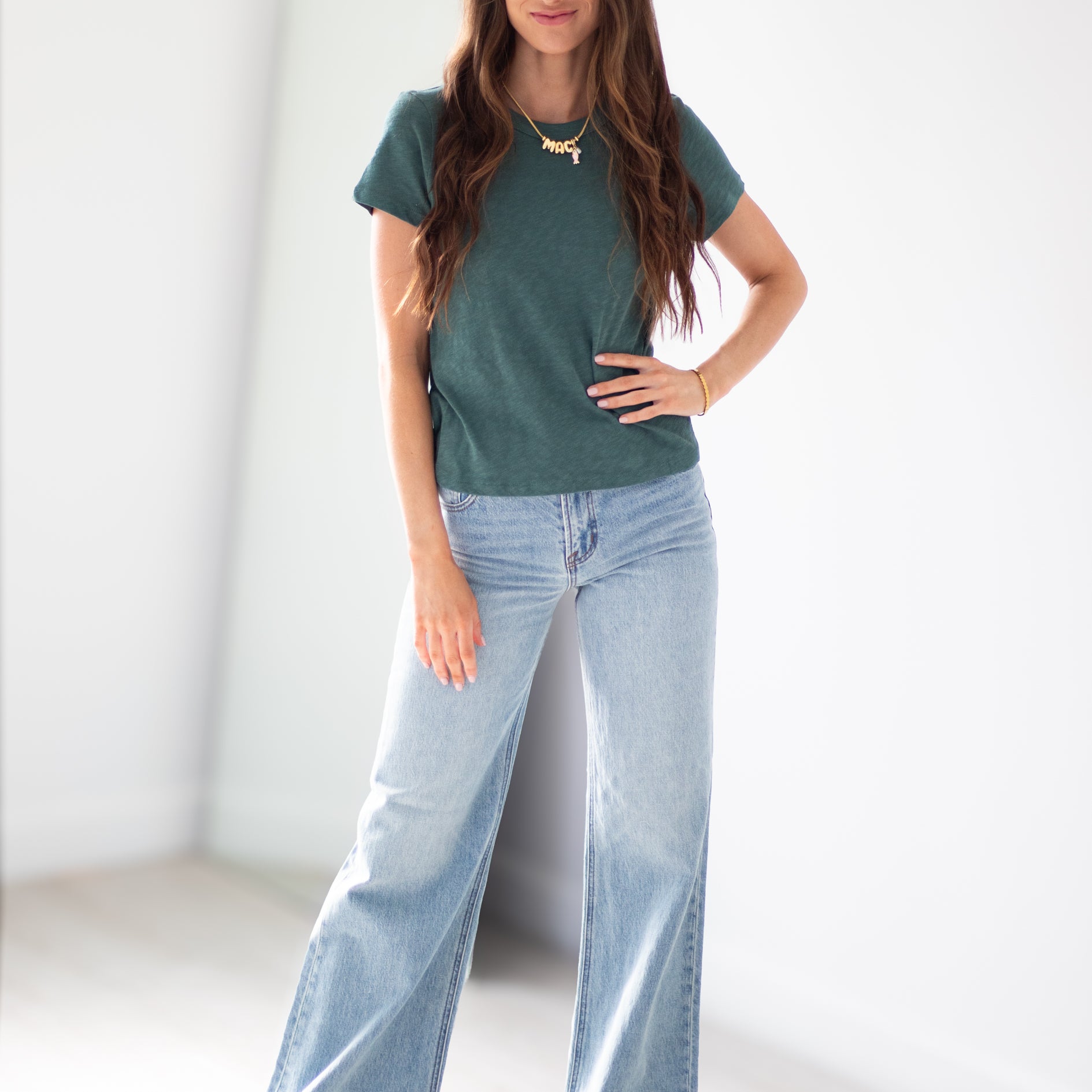 A woman modeling a dark green short-sleeve knit t-shirt and light-wash wide-leg denim jeans, standing in a bright, minimalist white room.